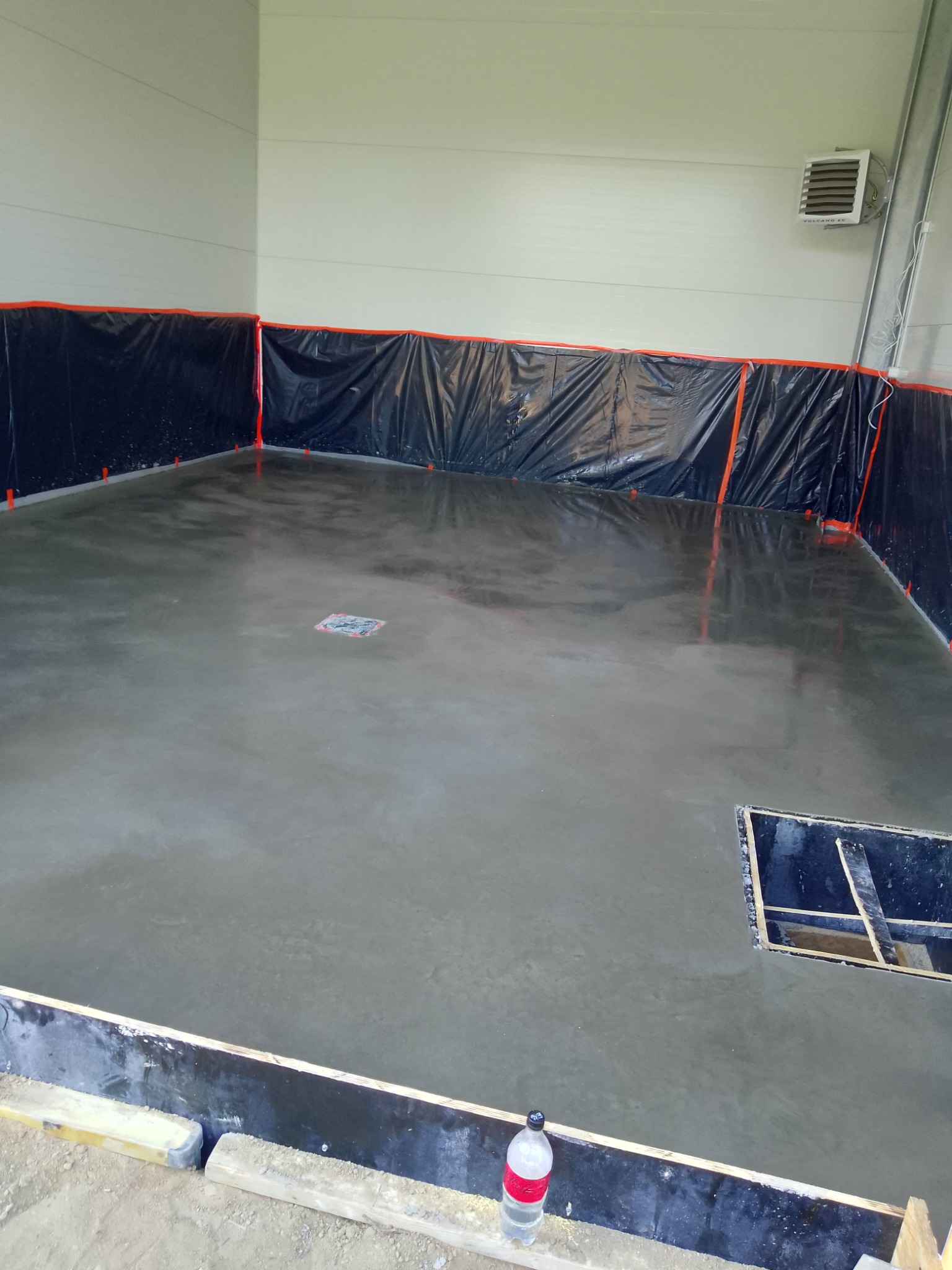 Świeżo wylana, gładka posadzka betonowa w hali, zabezpieczona folią przy ścianach. Widoczny odpływ liniowy i fragment szalunku. Butelka z wodą w rogu.