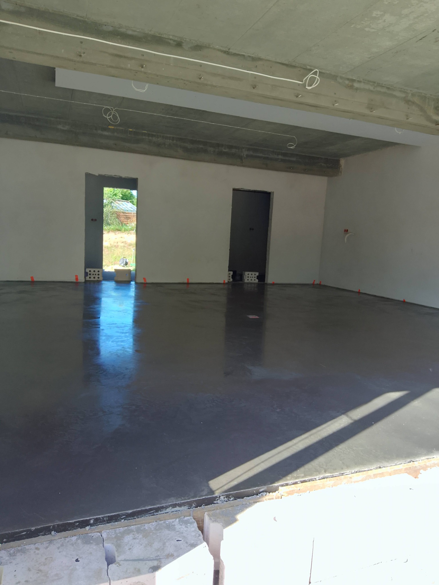 Wnętrze z wylaną, gładką posadzką. Widoczne pomarańczowe znaczniki na ścianach, surowy betonowy strop z instalacją elektryczną. Dwa otwory drzwiowe w tle.