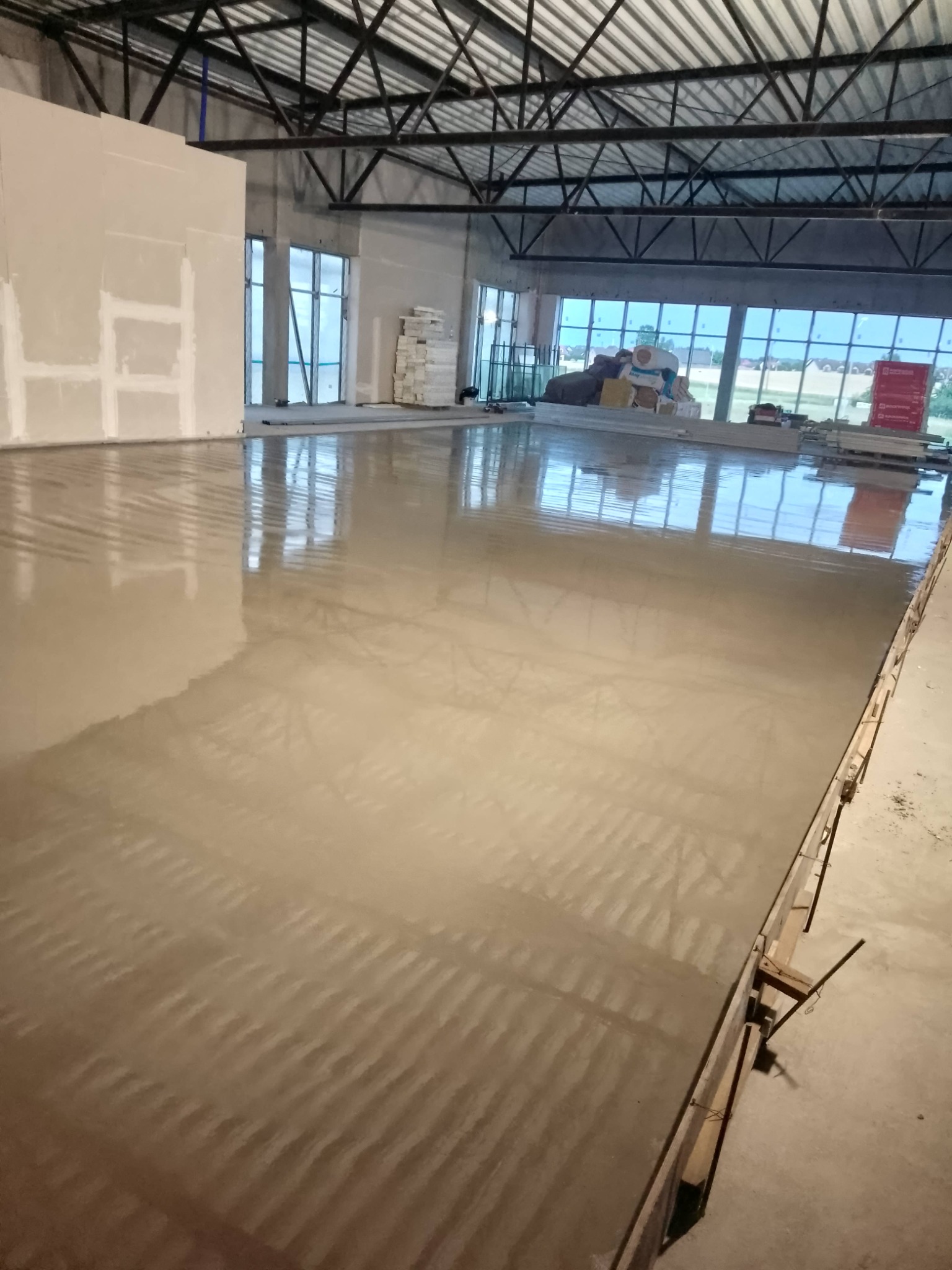 Świeżo wylana, mokra posadzka przemysłowa w hali z widocznymi oknami i materiałami budowlanymi. Elementy szalunku w prawym dolnym rogu.