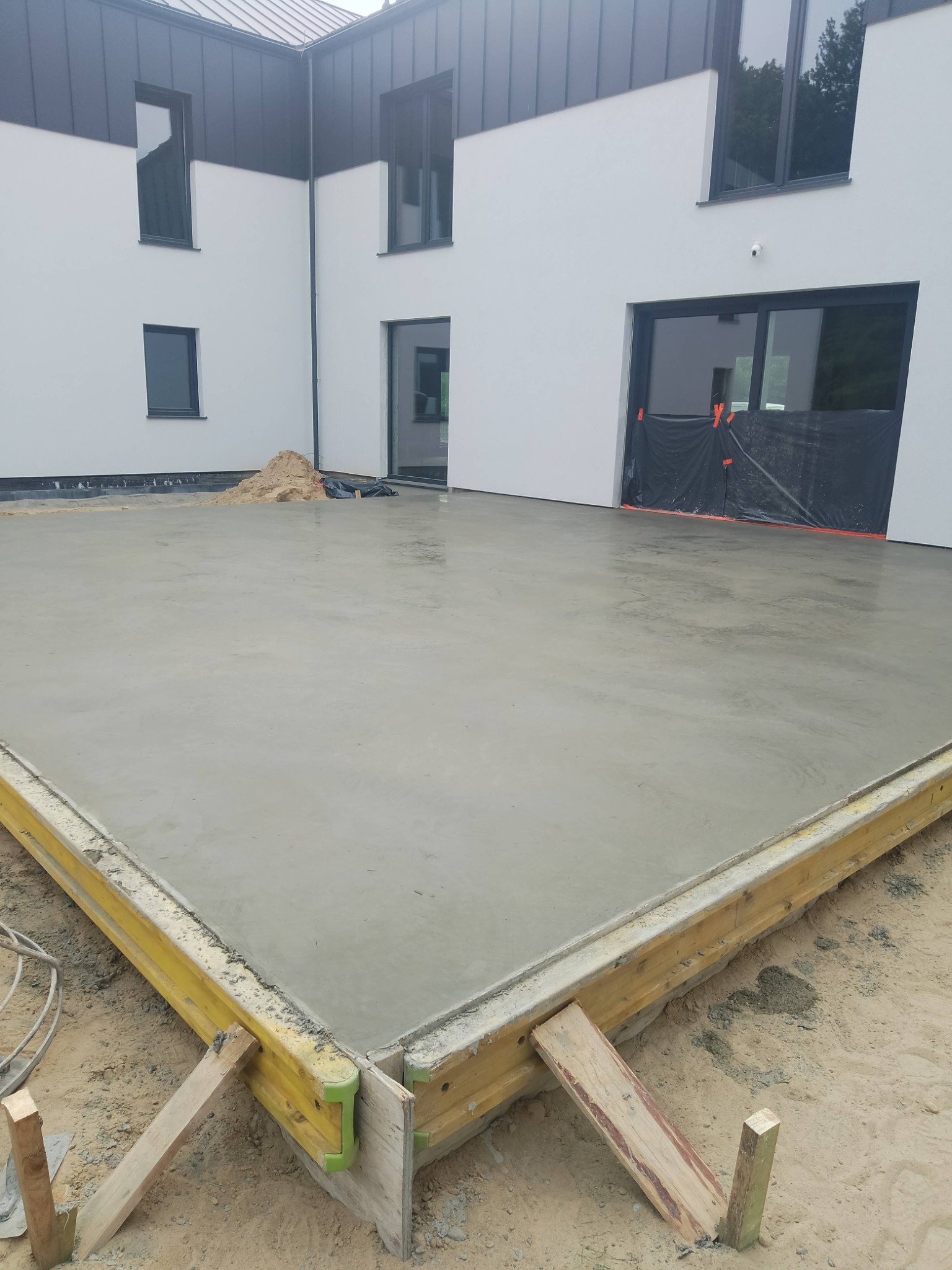 Świeżo wylana, gładka betonowa posadzka w szalunku, z widocznym fragmentem nowoczesnego domu z czarnymi oknami i dachem. Etap realizacji.