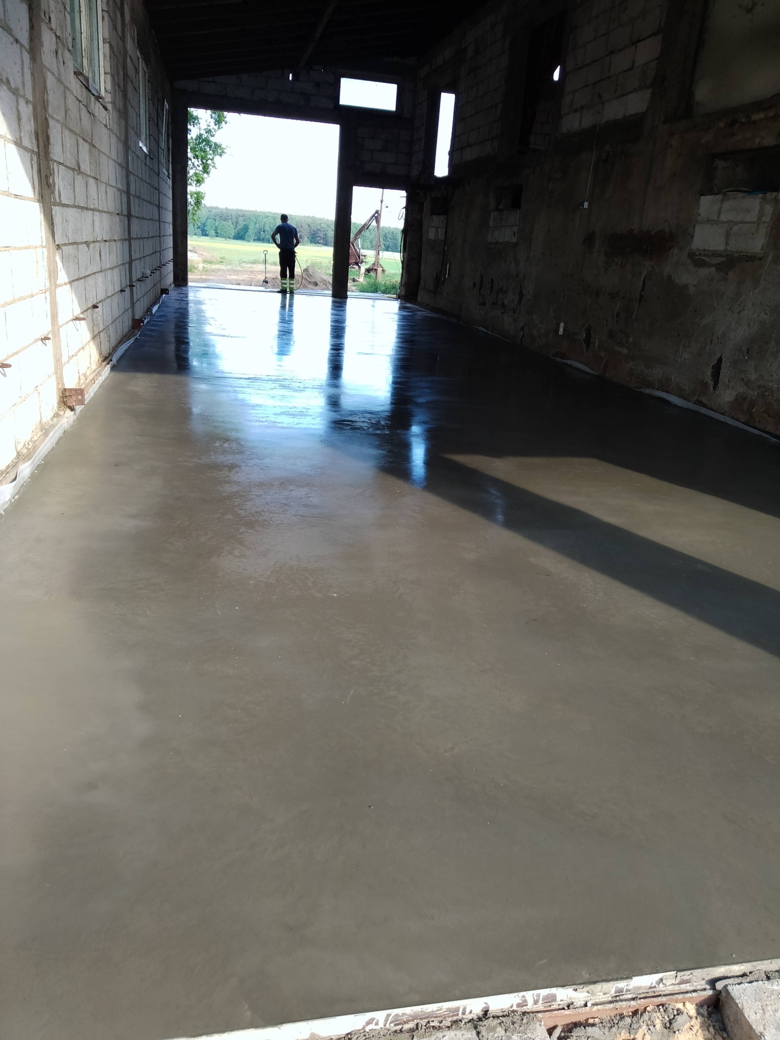 Świeżo wylana, gładka posadzka w budynku gospodarczym z widoczną osobą w oddali, nawadniającą powierzchnię. Widoczne ściany z cegły i bloczków.