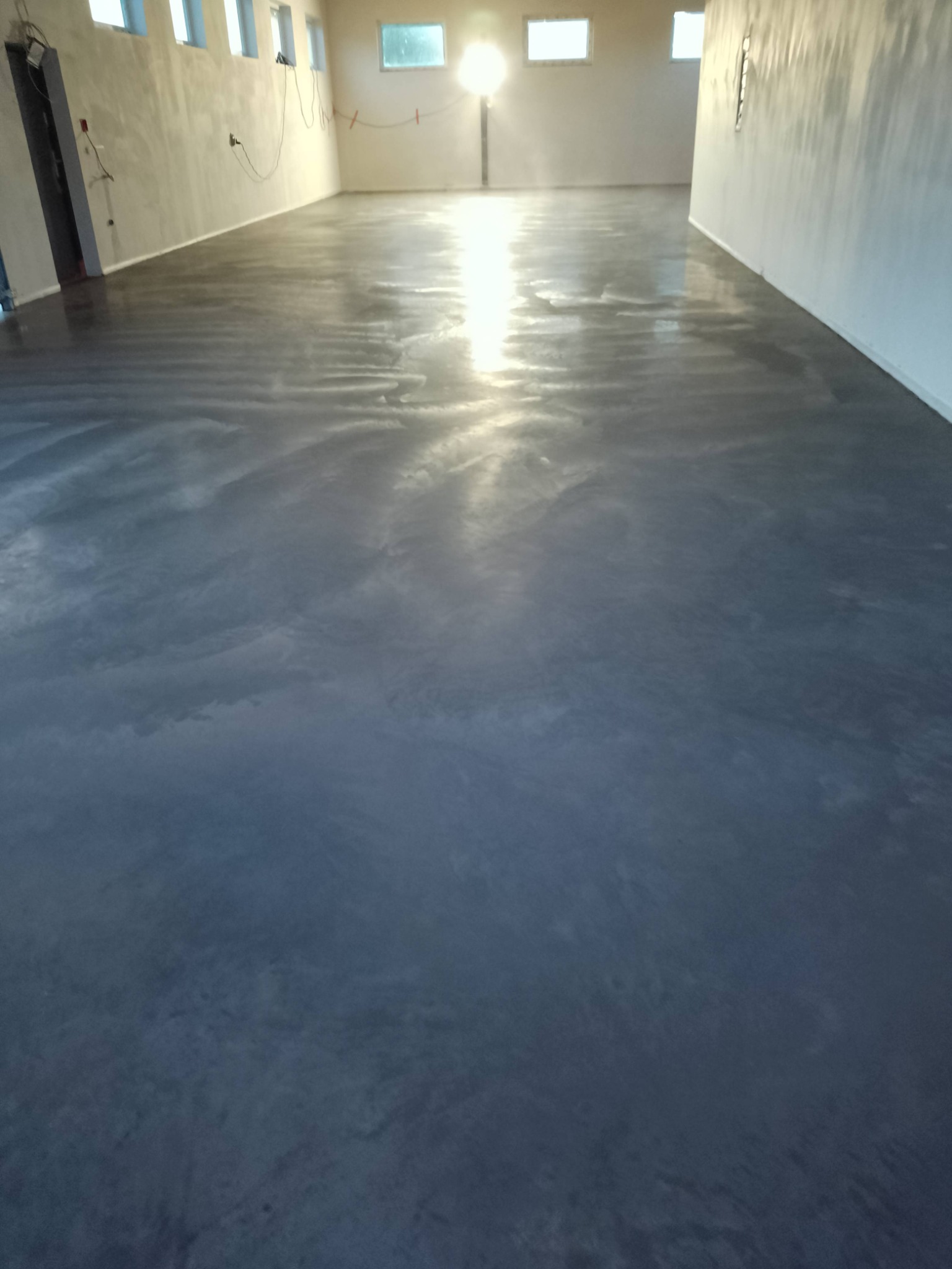 Błyszcząca, szara posadzka w hali przemysłowej z oknami, odbijająca światło słoneczne. Widoczne ślady po procesie wygładzania betonu.