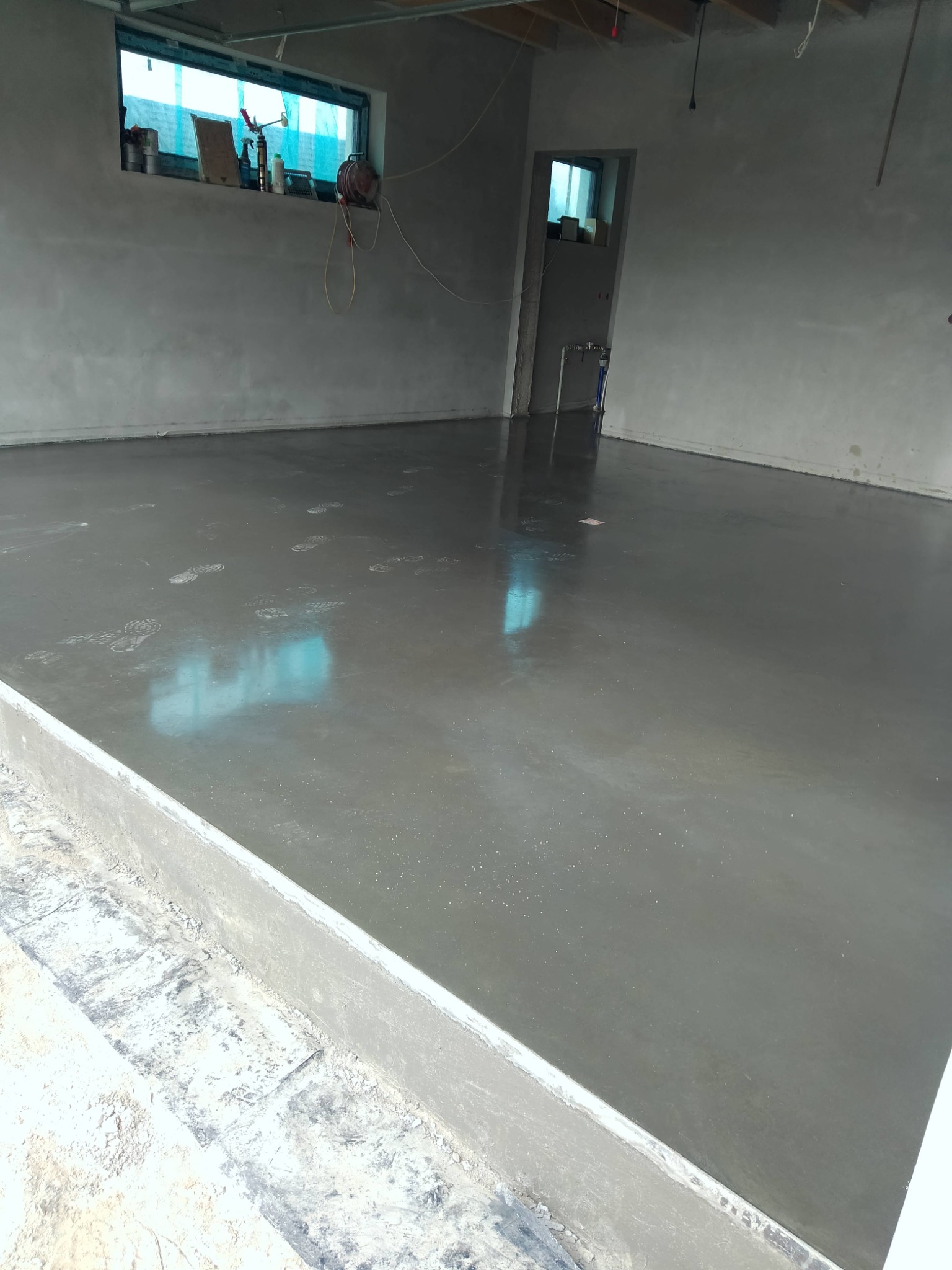 Świeżo wylana, gładka posadzka betonowa w szarym pomieszczeniu z oknem i śladami stóp. Widoczne narzędzia i materiały budowlane w tle.