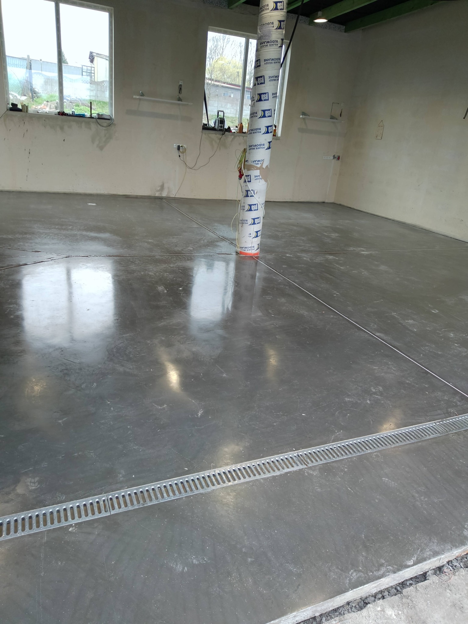 Wypoziomowana, szara posadzka przemysłowa w surowym wnętrzu, z widocznym odpływem liniowym i zabezpieczonym słupem. W tle okna i ściany przygotowane do wykończenia.