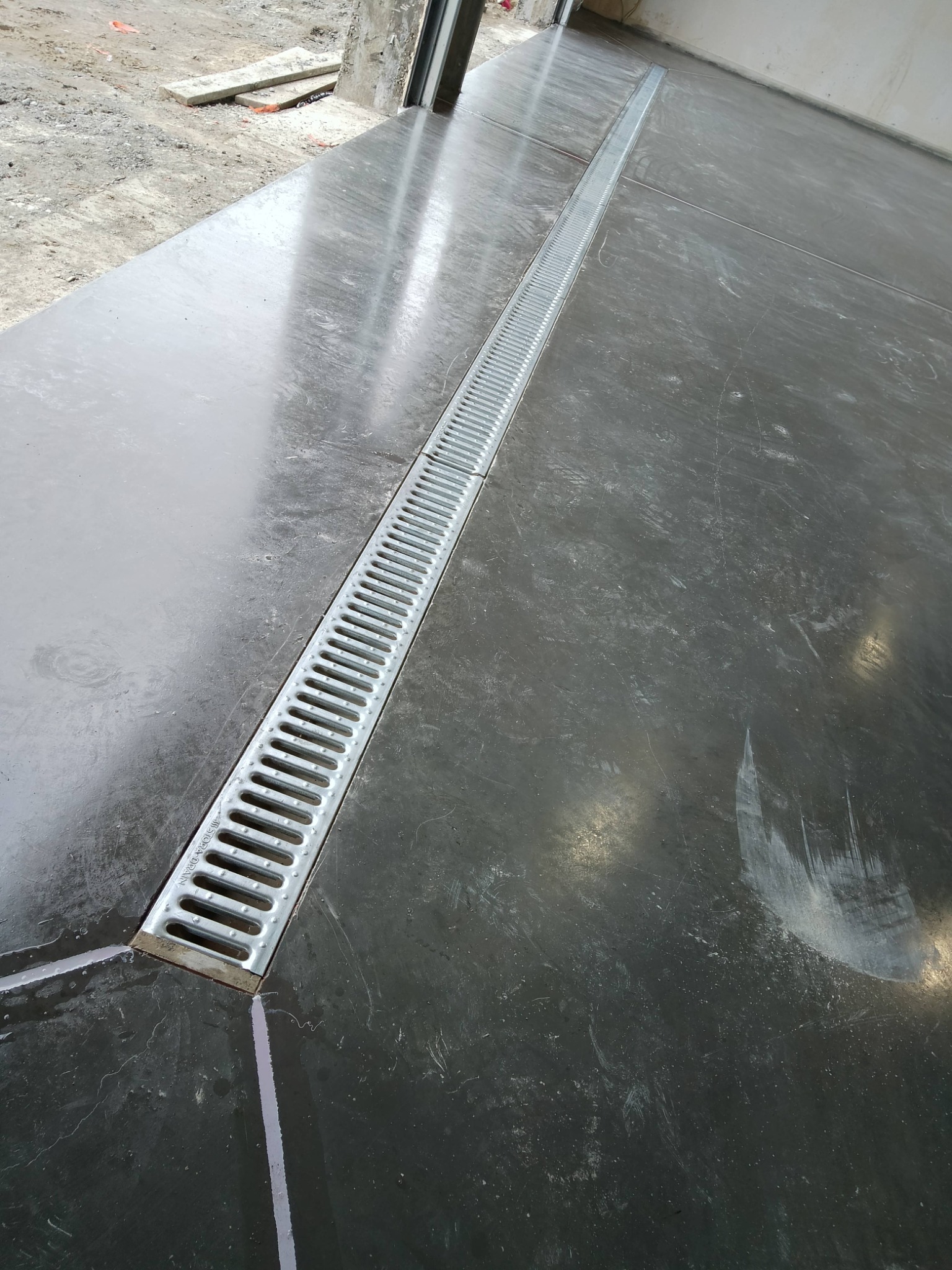 Szara, gładka posadzka z metalowym odpływem liniowym, ułożona wzdłuż przesuwanych drzwi balkonowych. Widoczne refleksy światła i surowe wykończenie.