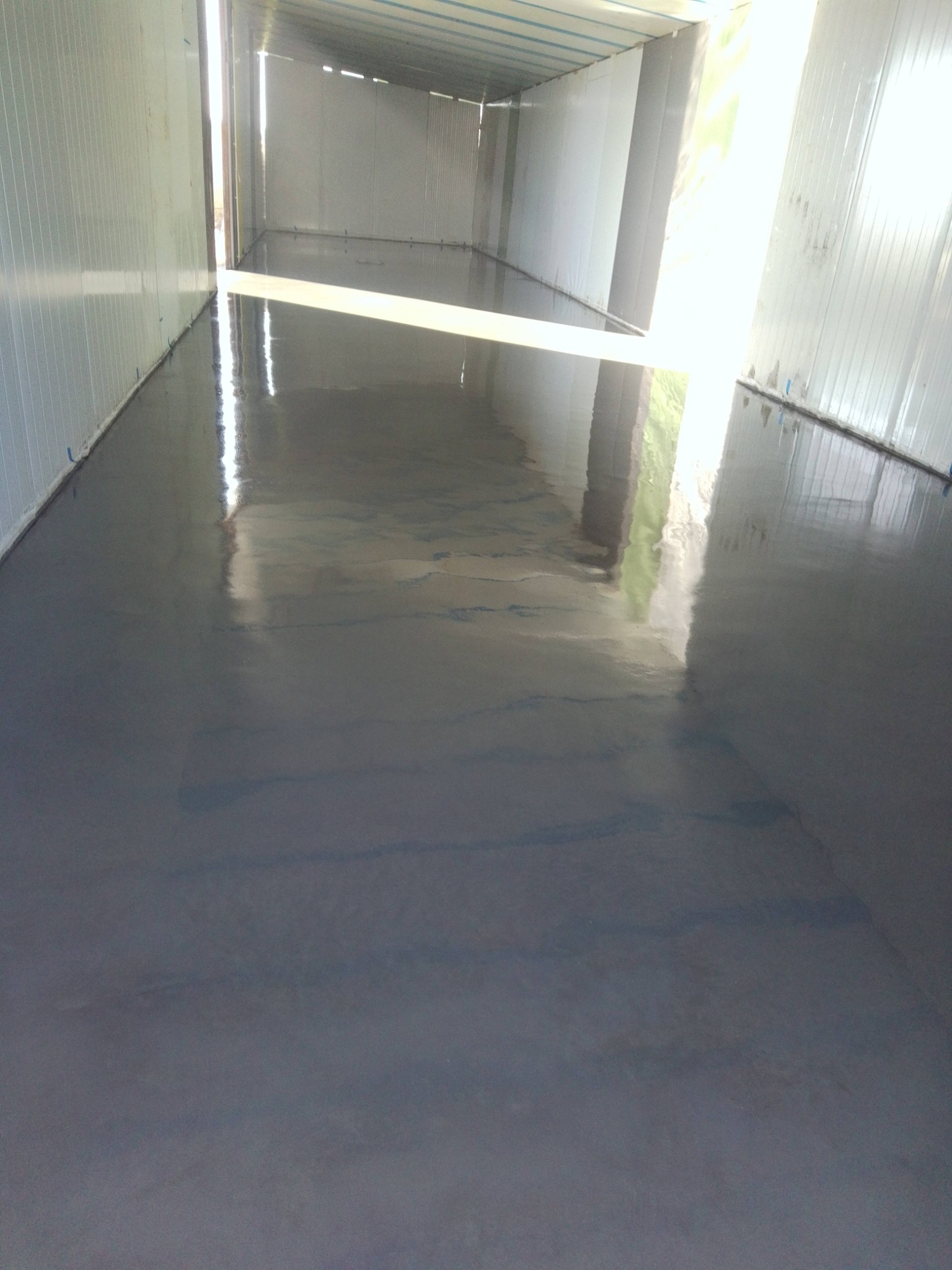 Błyszcząca, szara posadzka w hali przemysłowej z panelami ściennymi. Światło dzienne wpadające przez otwarte wrota. Widok z perspektywy podłogi.