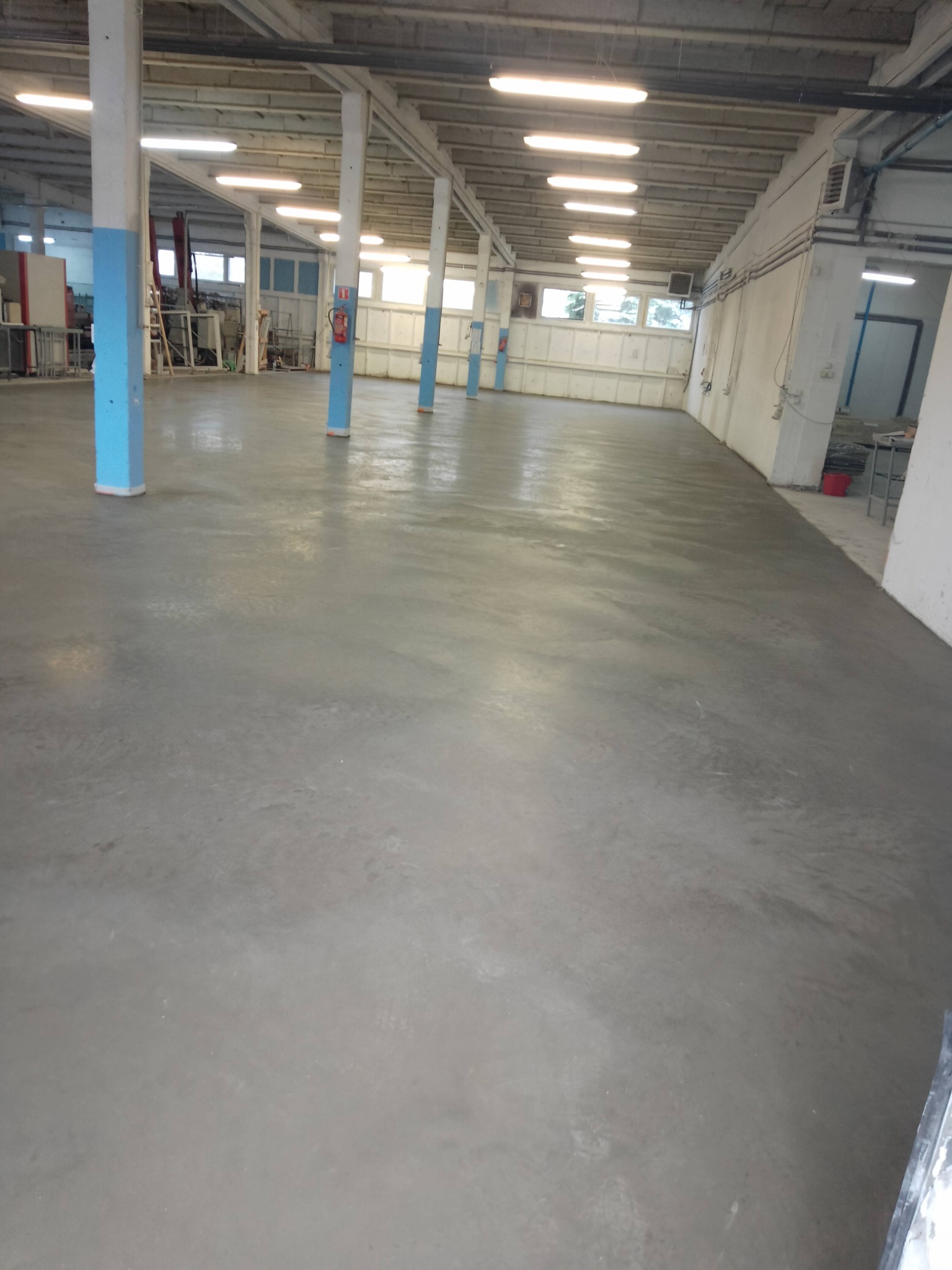 Gładka, szara posadzka przemysłowa w hali z niebieskimi słupami i rzędami lamp. Widoczne okna w tle i fragmenty wyposażenia. Świeża, równa powierzchnia.