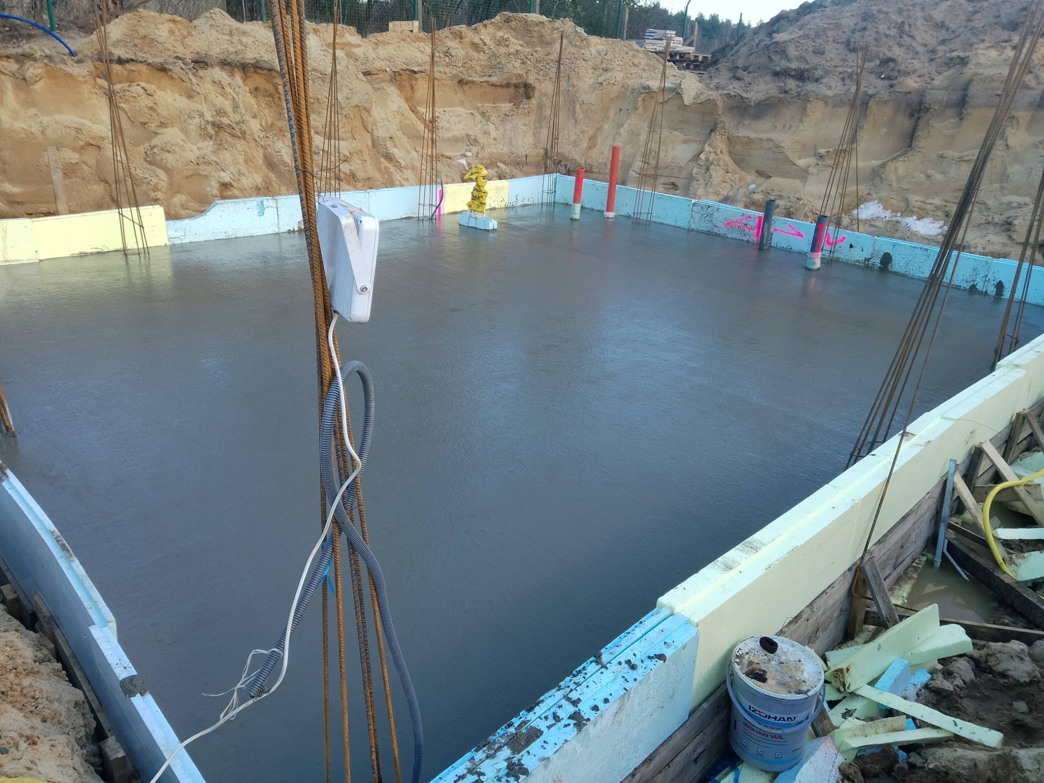 Wylany beton na przygotowany fundament z izolacją, widoczne wystające pręty zbrojeniowe i rury instalacyjne. Na pierwszym planie wiadro z farbą.