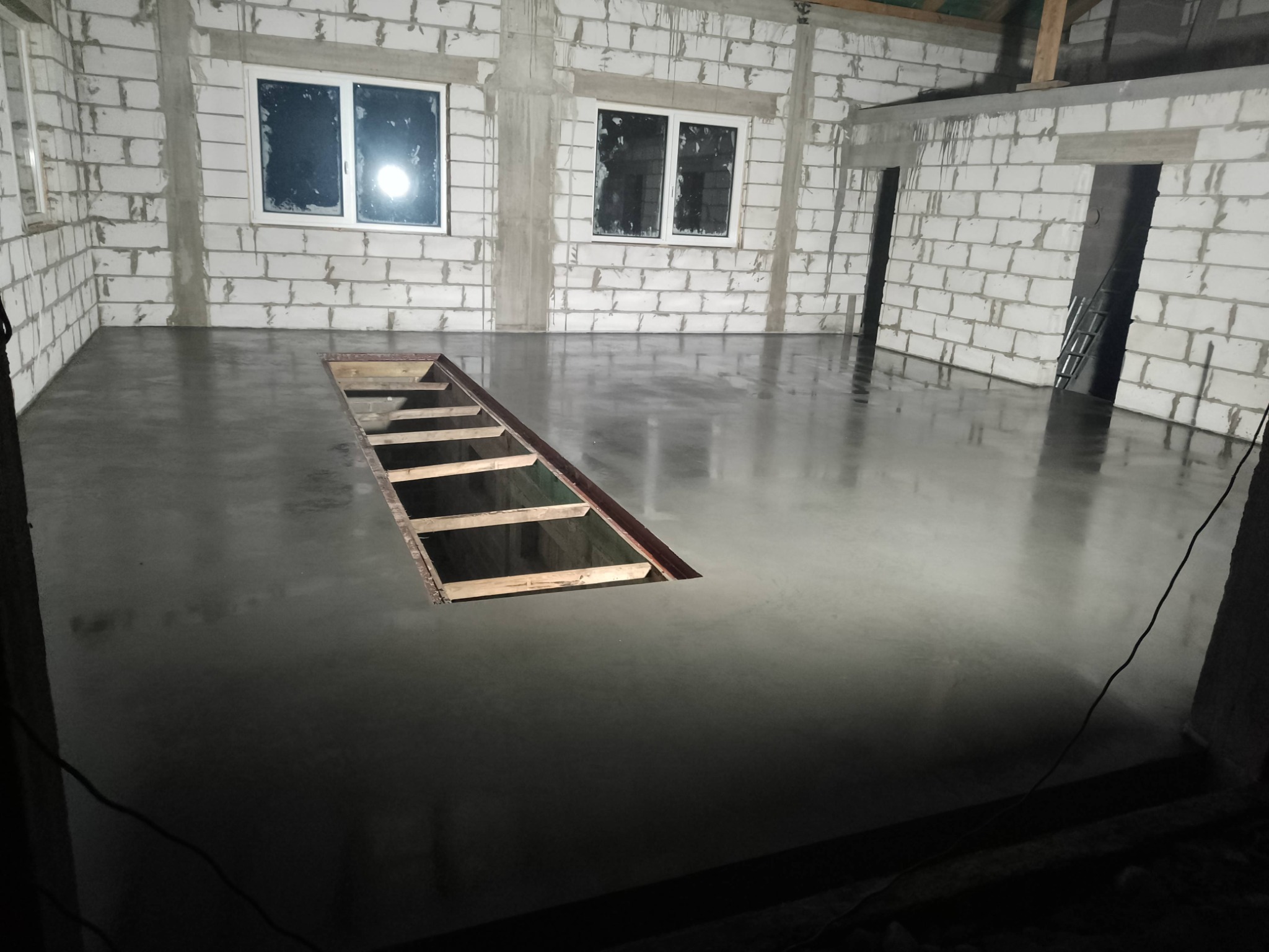 Świeżo wylana, gładka posadzka w surowym wnętrzu z białego bloczka; widoczne okna, otwór w podłodze z drewnianą konstrukcją, odblaski światła na mokrej powierzchni.