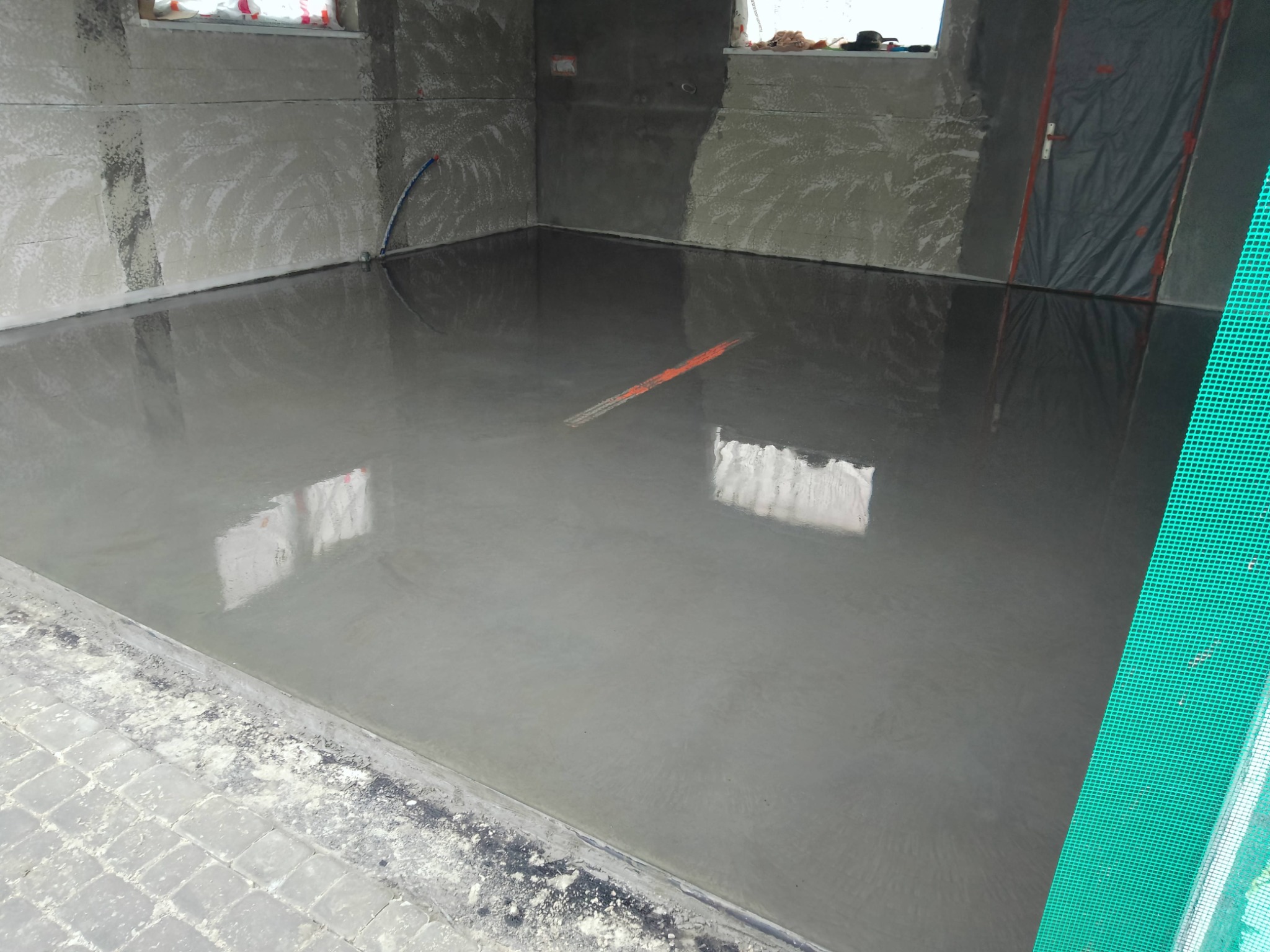 Świeżo wylana, gładka posadzka betonowa w surowym wnętrzu, z widoczną łatą pomarańczową i zabezpieczoną folią ścianą. Widoczne ślady po łatach na ścianach.