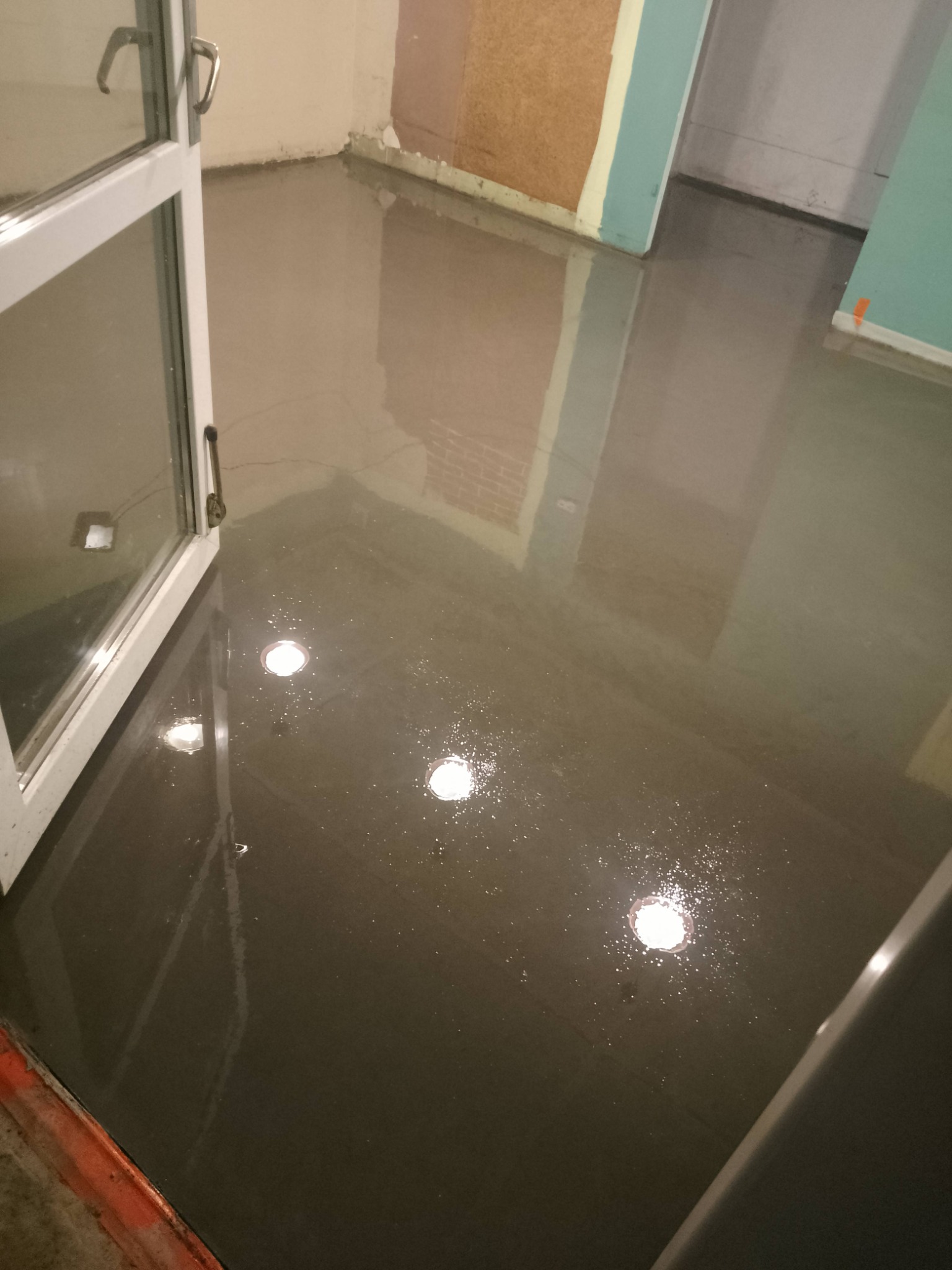 Świeżo wylana, mokra posadzka w pomieszczeniu z otwartym oknem. Widoczne odbicia światła w mokrej powierzchni. Przy ścianach widoczne zabezpieczenia.