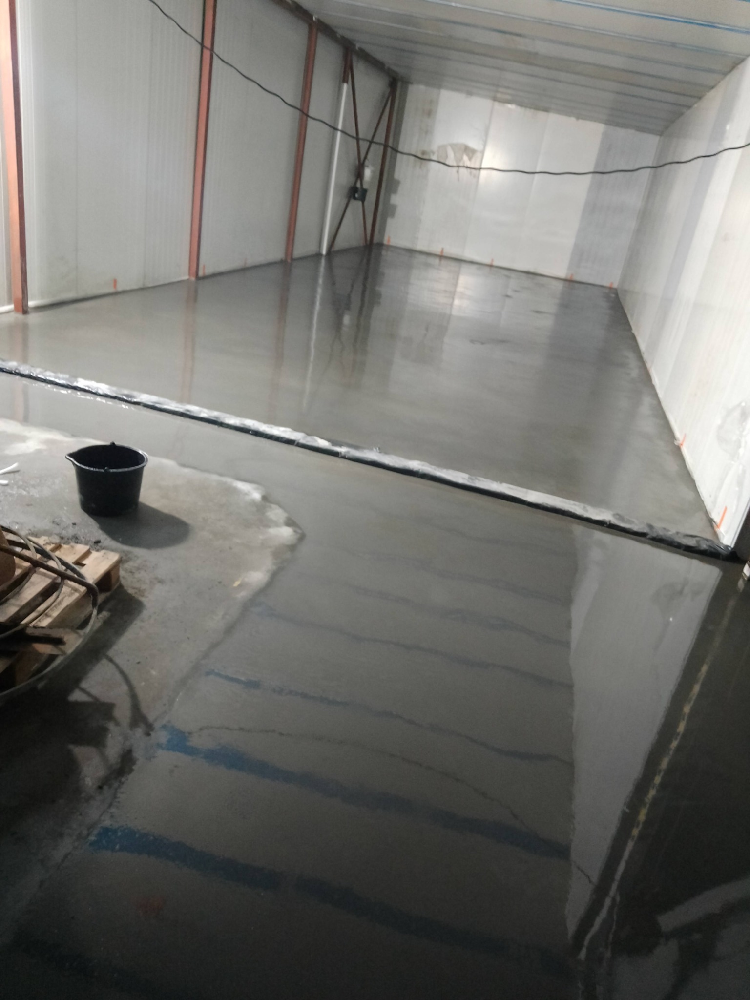 Świeżo wylana, gładka posadzka przemysłowa w hali z blachy trapezowej, z widocznym czarnym wiadrem i narzędziami po lewej stronie. Podłoga mokra, odbija światło.