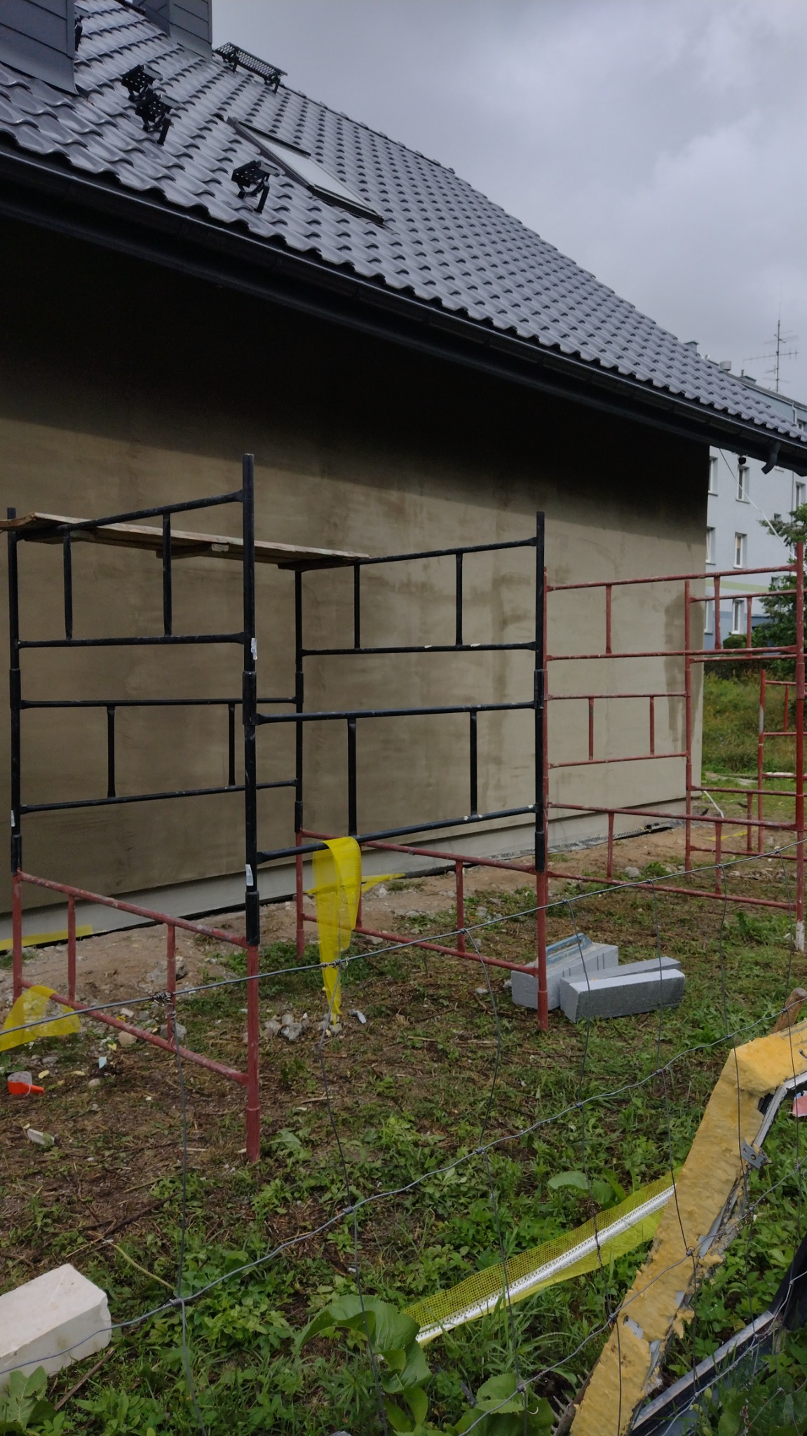 Ściana budynku w trakcie prac wykończeniowych, widoczne rusztowania, ocieplenie w siatce, dach z czarnej dachówki, teren zarośnięty trawą.