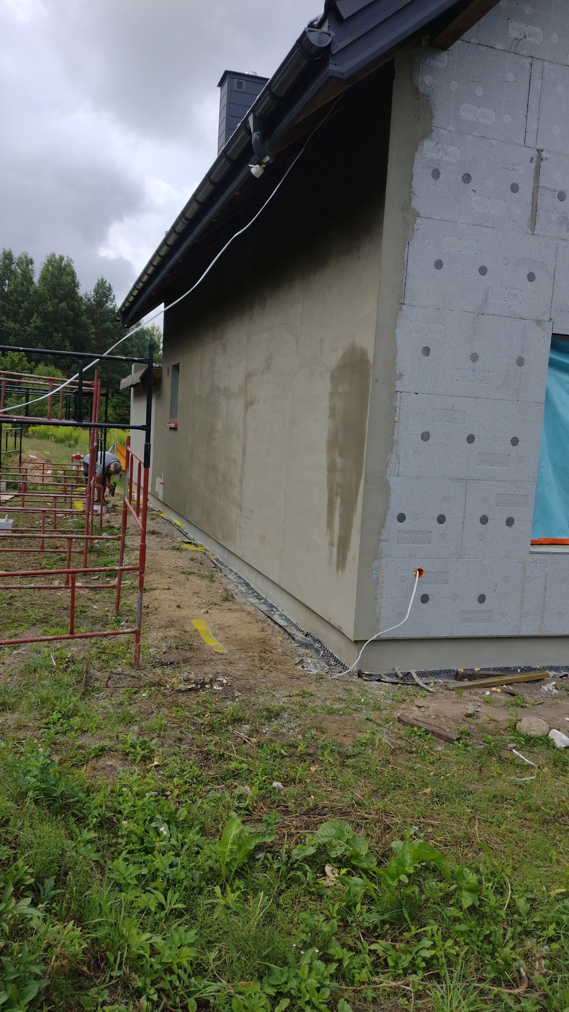 Fragment domu w trakcie prac wykończeniowych: ocieplona ściana z szarymi płytami styropianowymi i fragment ściany z nałożonym szarym tynkiem, widoczne rusztowanie i kabel elektryczny wystający ze...