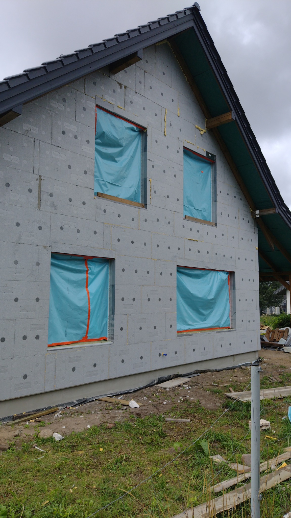 Ściana domu w trakcie ocieplania, pokryta szarymi płytami izolacyjnymi z widocznymi okrągłymi zagłębieniami, okna zabezpieczone niebieską folią, widoczne fragmenty pianki montażowej w szczelinach.