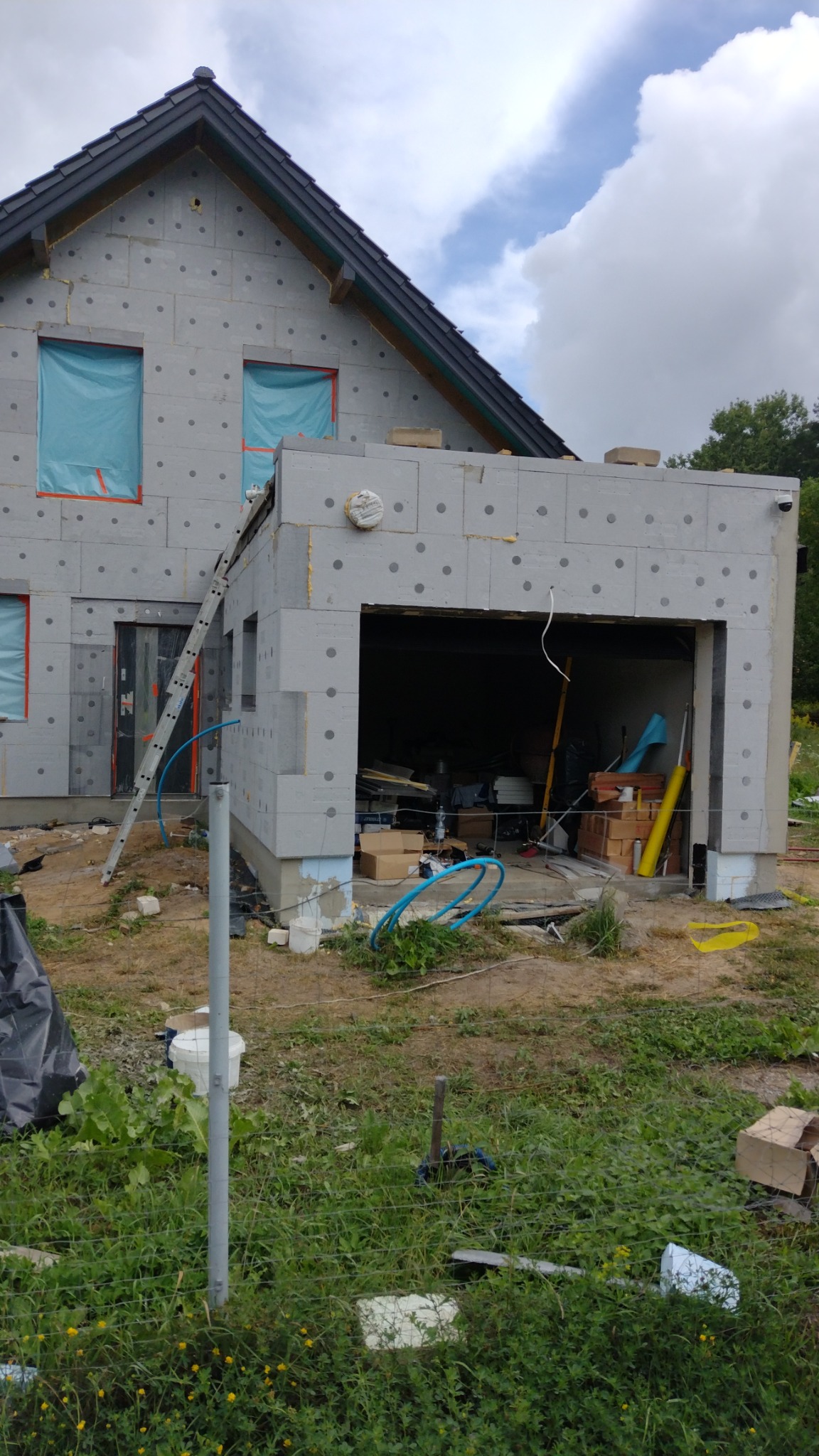 Szary dom w trakcie budowy z widocznym ociepleniem ścian zewnętrznych, zabezpieczonymi oknami folią, drabina oparta o ścianę oraz otwarty garaż z widocznymi materiałami budowlanymi w środku.