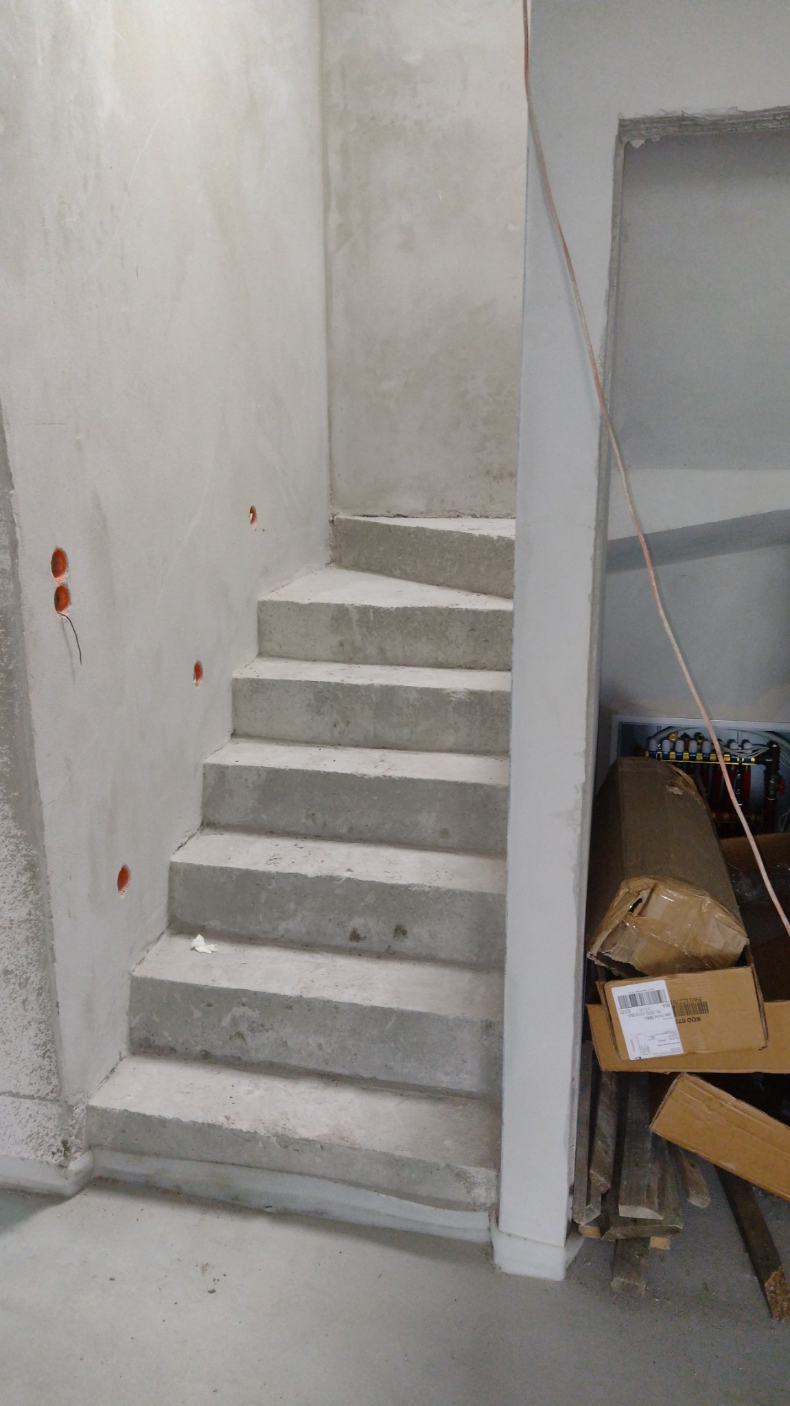 Surowe betonowe schody spiralne w trakcie budowy, z widocznymi otworami pod instalacje elektryczne w ścianie i kartonami w tle.