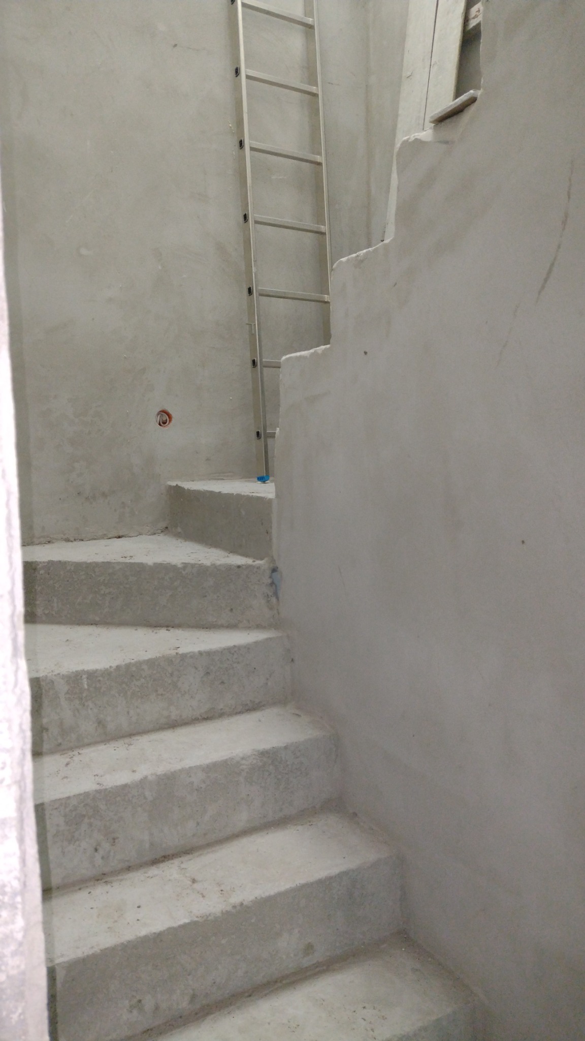 Surowe, betonowe schody wewnętrzne w trakcie budowy, z aluminiową drabiną opartą o ścianę, widoczne ślady po szalunku na ścianach.