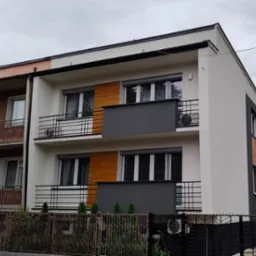 Dwupiętrowy budynek z balkonami i zamontowanymi klimatyzatorami na ścianach zewnętrznych, widziany zza ogrodzenia.