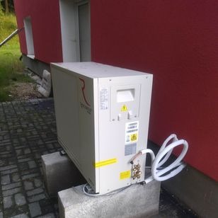 Biała jednostka zewnętrzna pompy ciepła ustawiona na betonowych bloczkach przy czerwonej ścianie budynku. Widoczne rury przyłączeniowe i etykiety ostrzegawcze.