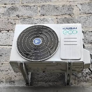 Zewnętrzna jednostka klimatyzatora Kaisai Eco zamontowana na szarej, ceglanej ścianie. Widoczne metalowe wsporniki mocujące urządzenie.