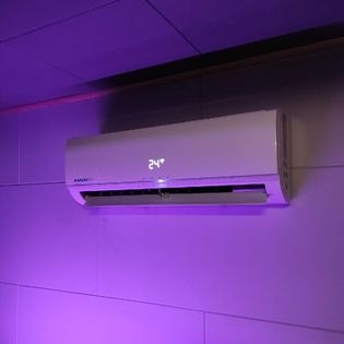 Biały klimatyzator ścienny z wyświetlaną temperaturą 24 stopnie Celsjusza, oświetlony fioletowym światłem, zamontowany na jasnej, panelowej ścianie.