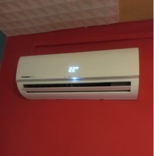 Biały klimatyzator Kaisai zamontowany na czerwonej ścianie z wyświetlaną temperaturą 22 stopnie Celsjusza, widoczny fragment sufitu z kasetonami.