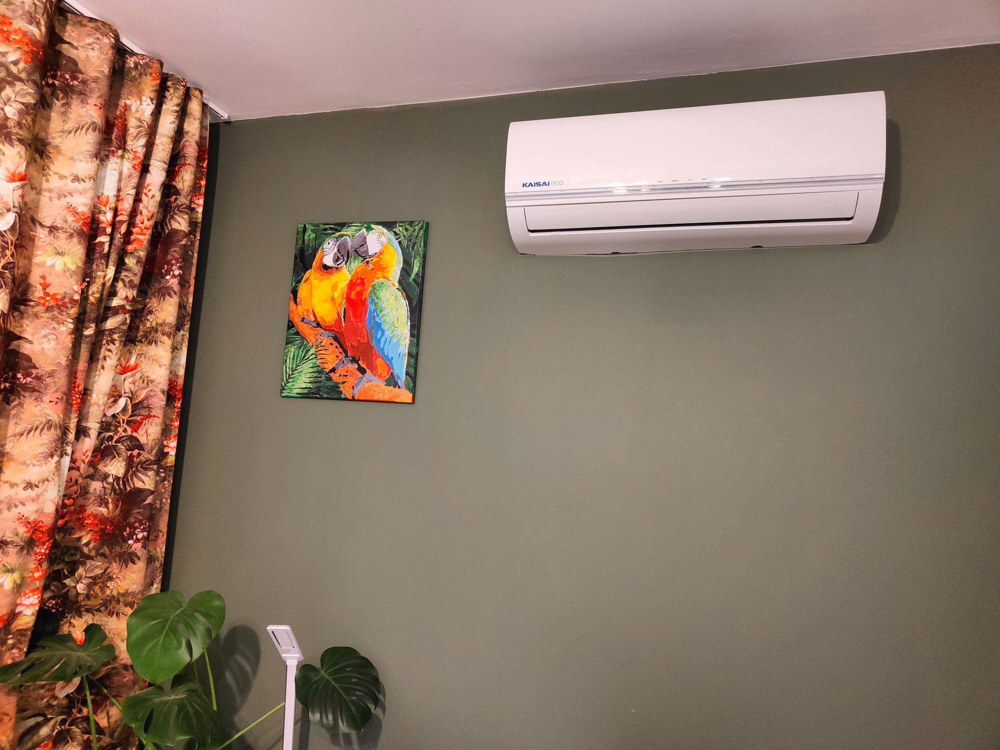 Biały klimatyzator marki Kaisai Eco zamontowany na oliwkowozielonej ścianie, obok obrazu z papugami i zasłony w kwiaty, z widoczną rośliną Monstera w rogu.