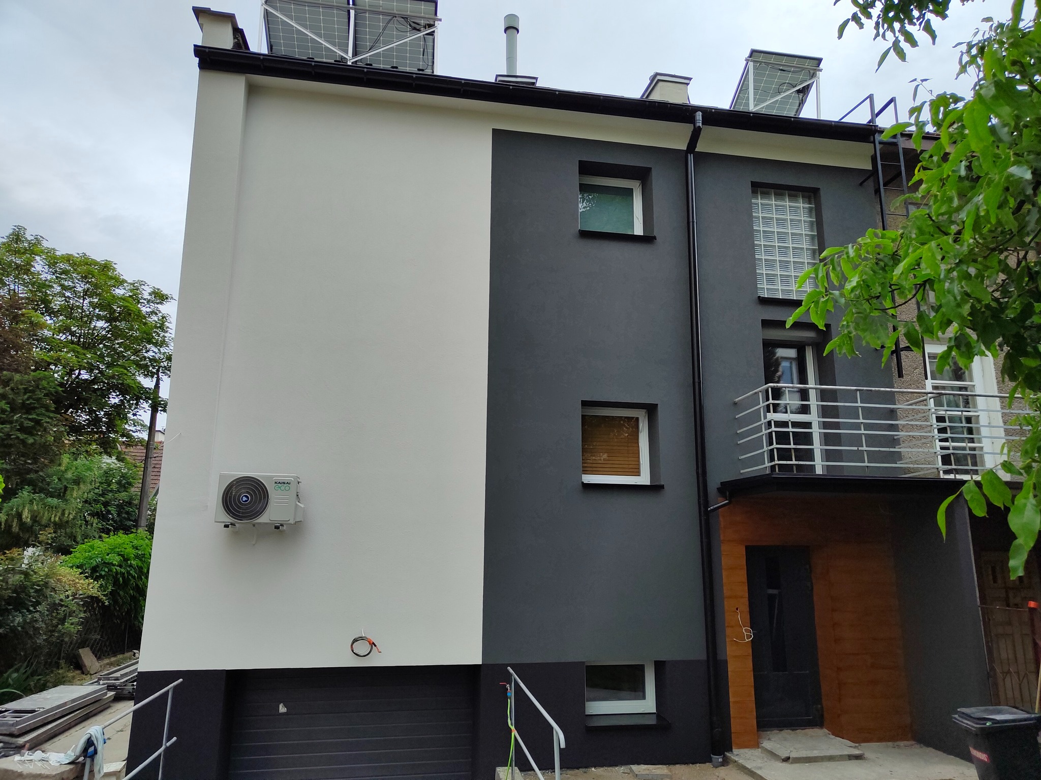 Dwukolorowy budynek z zamontowanym klimatyzatorem Kaisai Eco na białej ścianie, panele fotowoltaiczne na dachu i widoczne elementy instalacji elektrycznej przy klimatyzatorze.