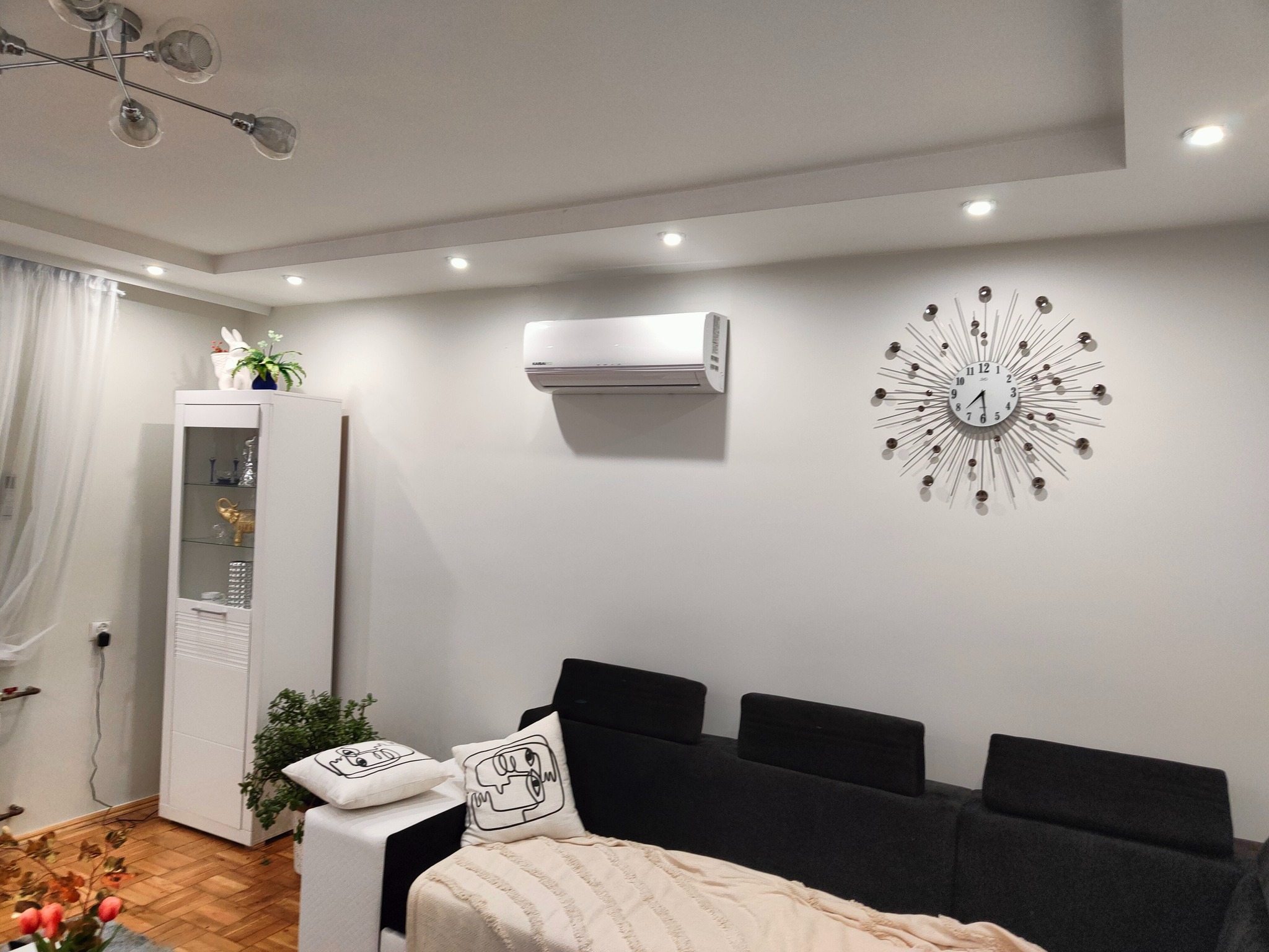 Nowoczesne wnętrze salonu z zamontowanym klimatyzatorem ściennym, dekoracyjnym zegarem słonecznym i minimalistyczną szafką witrynową.