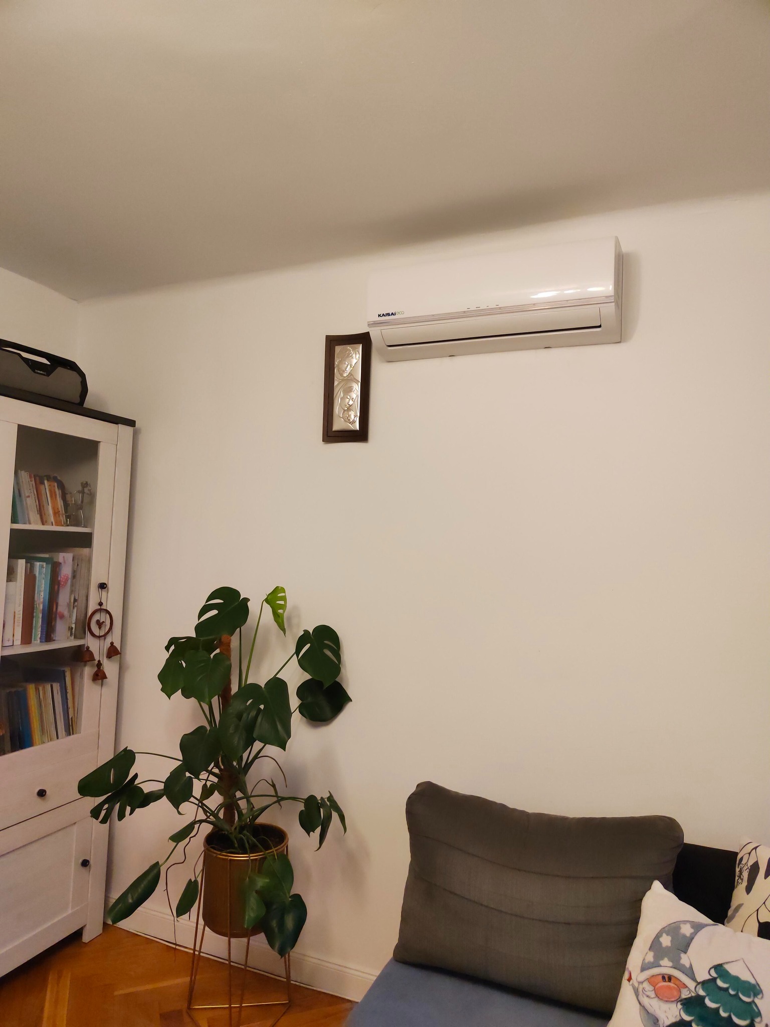 Biała jednostka klimatyzacji ściennej zamontowana w pokoju z minimalistycznym wystrojem, obok wiszącego obrazka z motywem rodzinnym, a poniżej stoi roślina Monstera w złotej doniczce na metalowym...