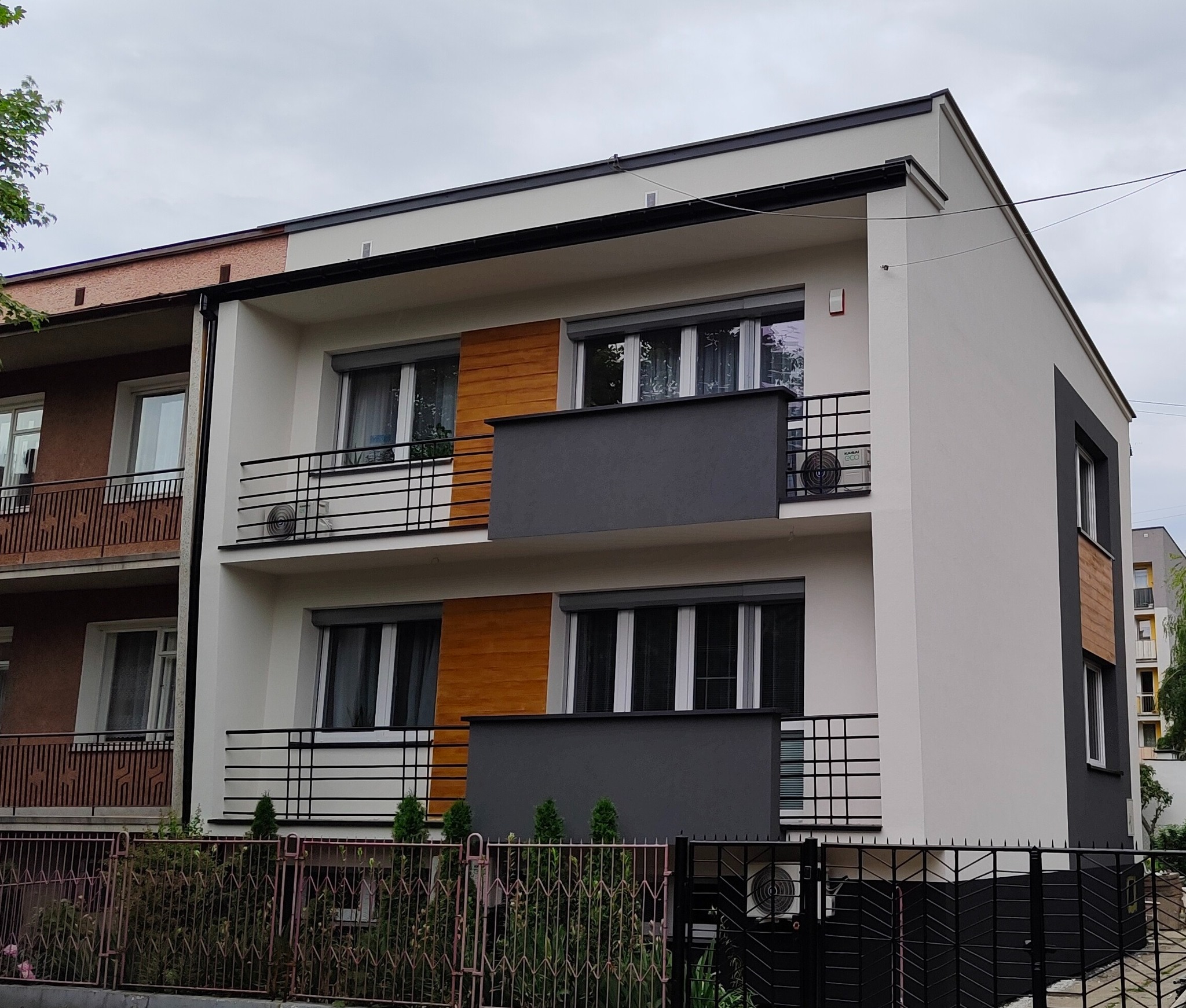 Dwupiętrowy budynek z balkonami i zamontowanymi klimatyzatorami na ścianach zewnętrznych, widziany zza ogrodzenia.