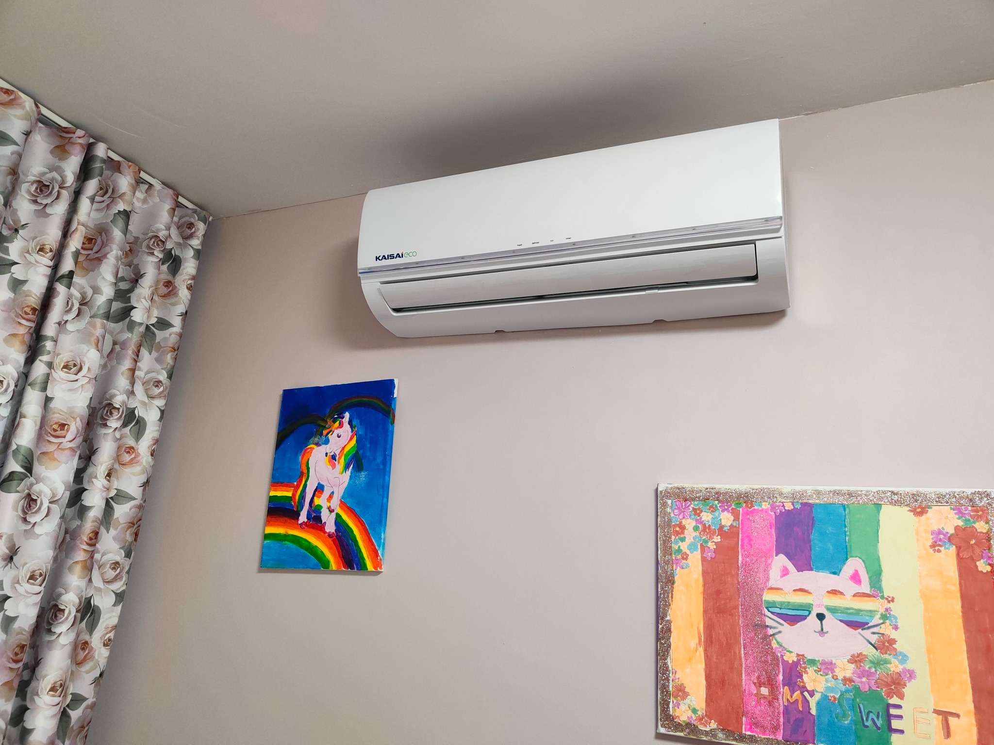 Biały klimatyzator Kaisai Eco zamontowany na ścianie w pokoju dziecięcym, obok zasłony w kwiaty i dwóch kolorowych obrazków namalowanych przez dzieci.