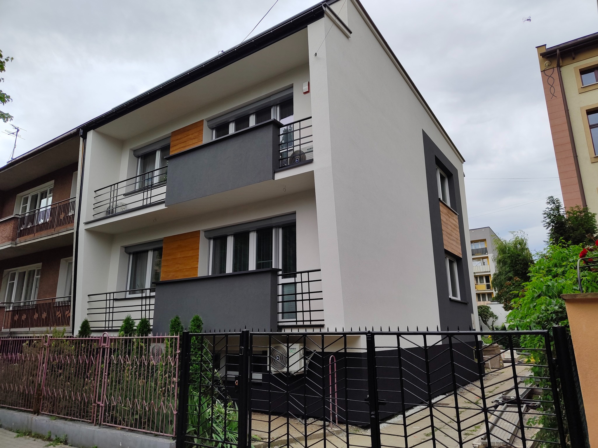 Nowoczesny, trzypiętrowy budynek z balkonami i klimatyzatorem na ścianie, widoczny zza czarnego, metalowego ogrodzenia.