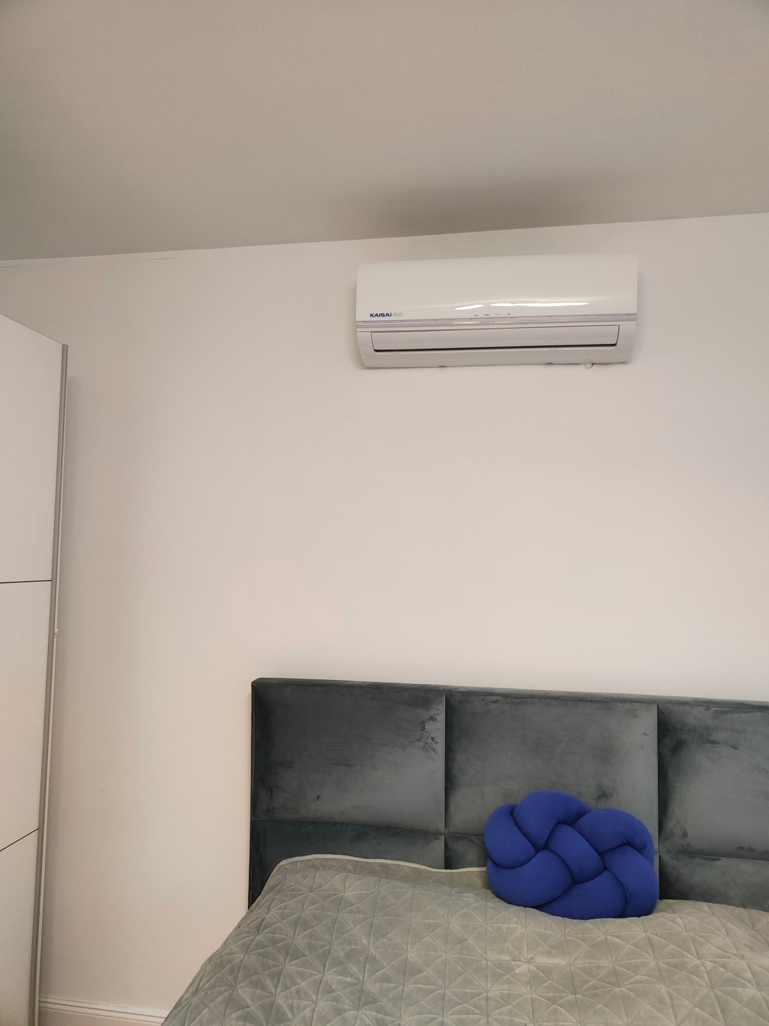 Biały klimatyzator ścienny marki KAISAI ECO zamontowany nad szarym wezgłowiem łóżka z ozdobną, niebieską poduszką w sypialni o minimalistycznym wystroju.