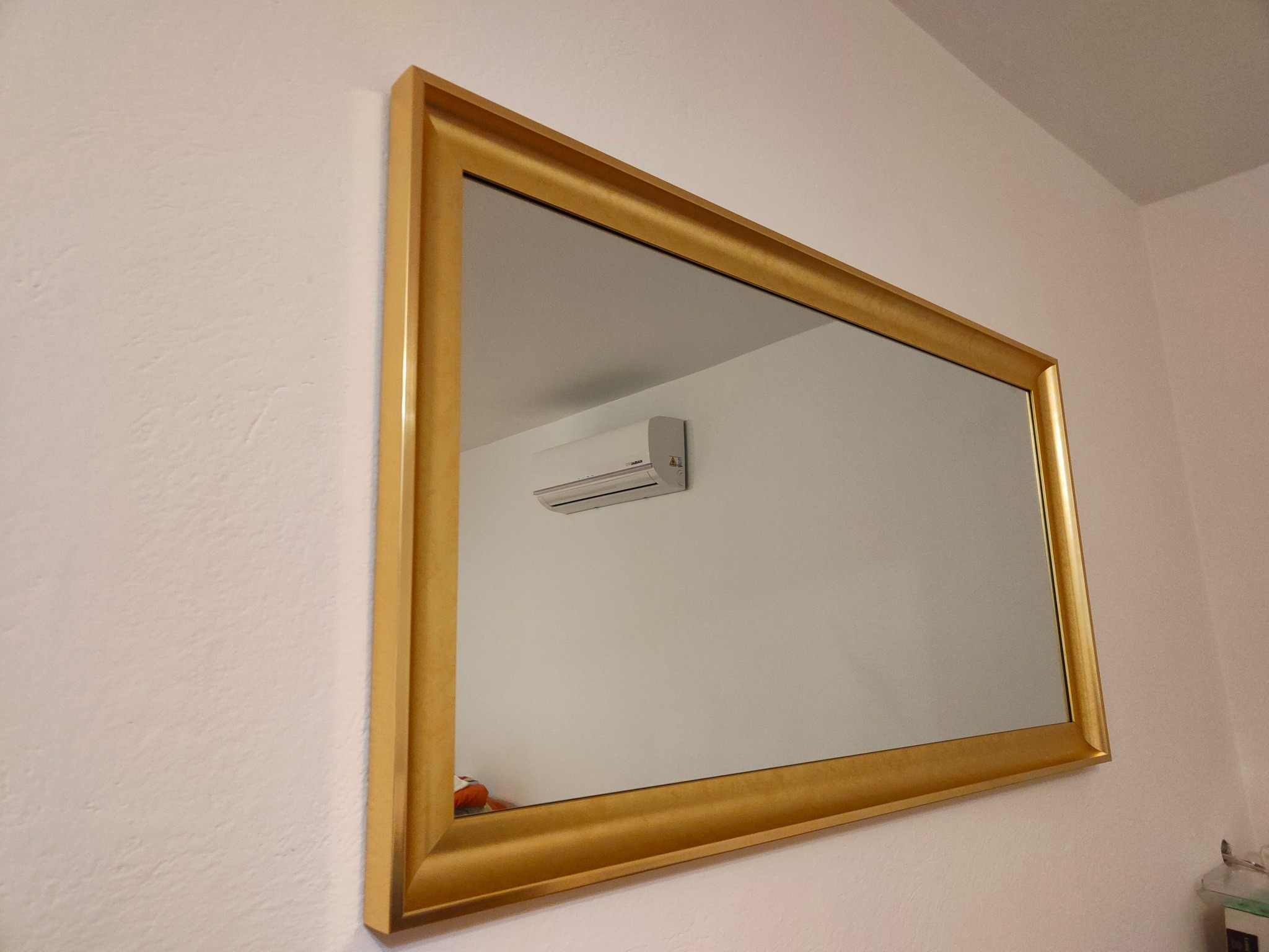 Złota rama lustra zawieszonego na ścianie, odbijająca klimatyzator na białej ścianie.