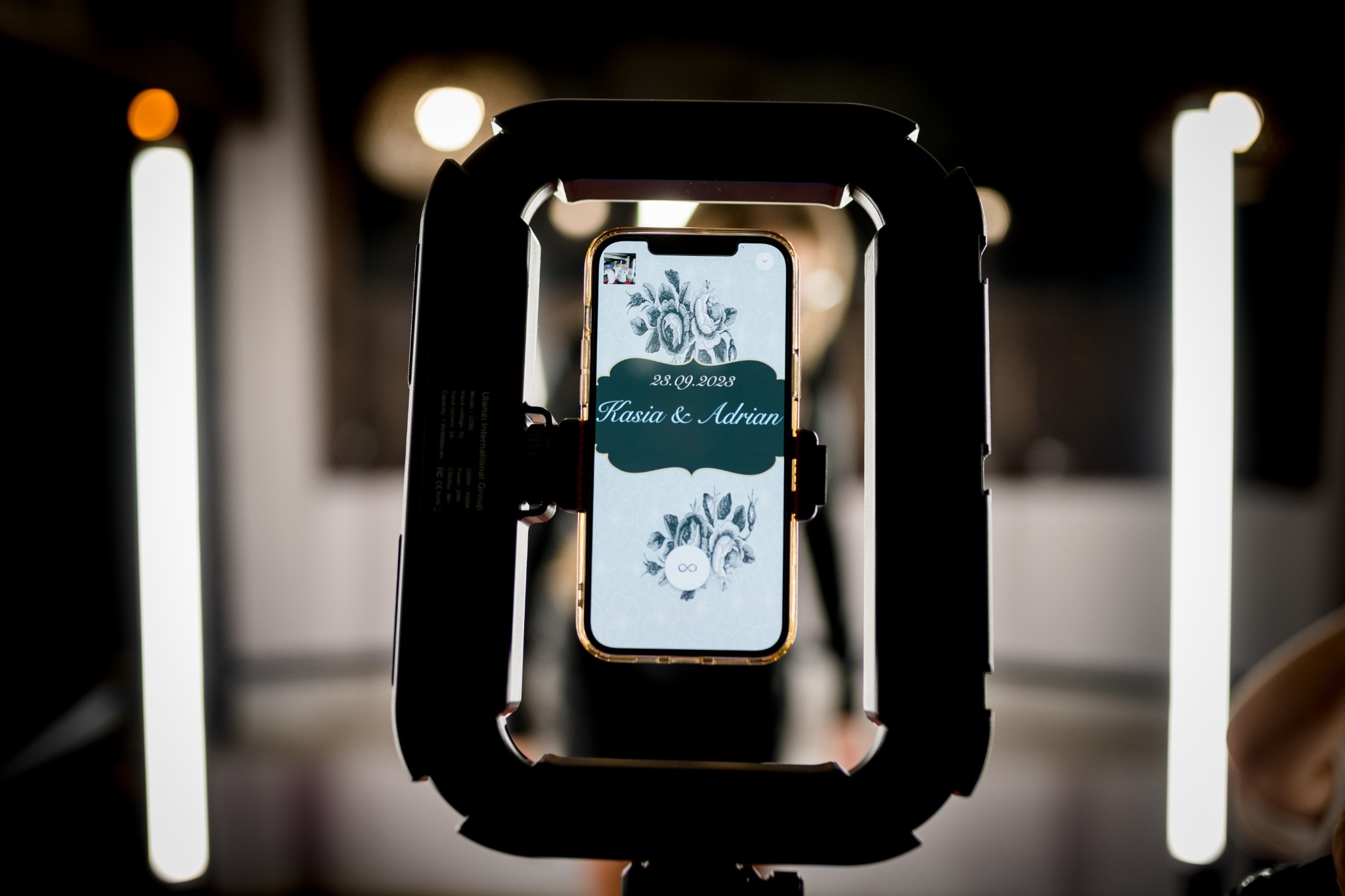 Wideobudka 360 z telefonem prezentującym spersonalizowany ekran powitalny z datą 23.09.2023 i imionami Kasia i Adrian, otoczona oświetleniem LED.