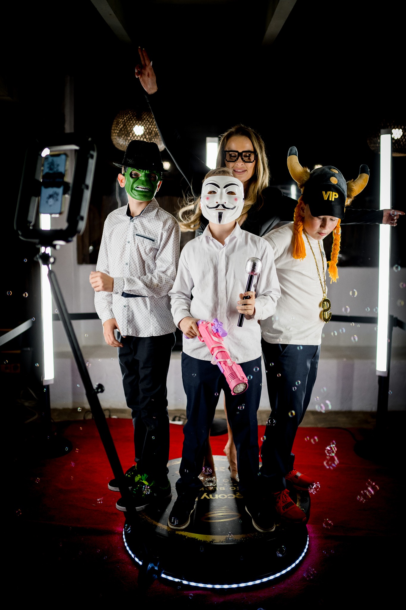 Uczestnicy w maskach i przebraniach pozują w wideobudce 360 otoczonej bańkami mydlanymi, na czerwonym dywanie, oświetlonej lampami LED.