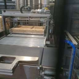 Andrei Sychou - Widok na automatyczną linię produkcyjną z podajnikiem kartonów, widoczne elementy mechaniczne, silniki, łańcuchy i przewody w hali przemysłowej.