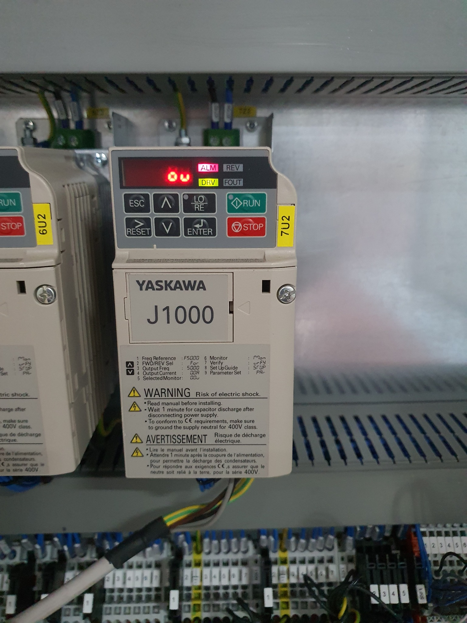 Panel sterowania Yaskawa J1000 z wyświetlaczem OU, przyciskami i okablowaniem, widoczne ostrzeżenie o ryzyku porażenia prądem.