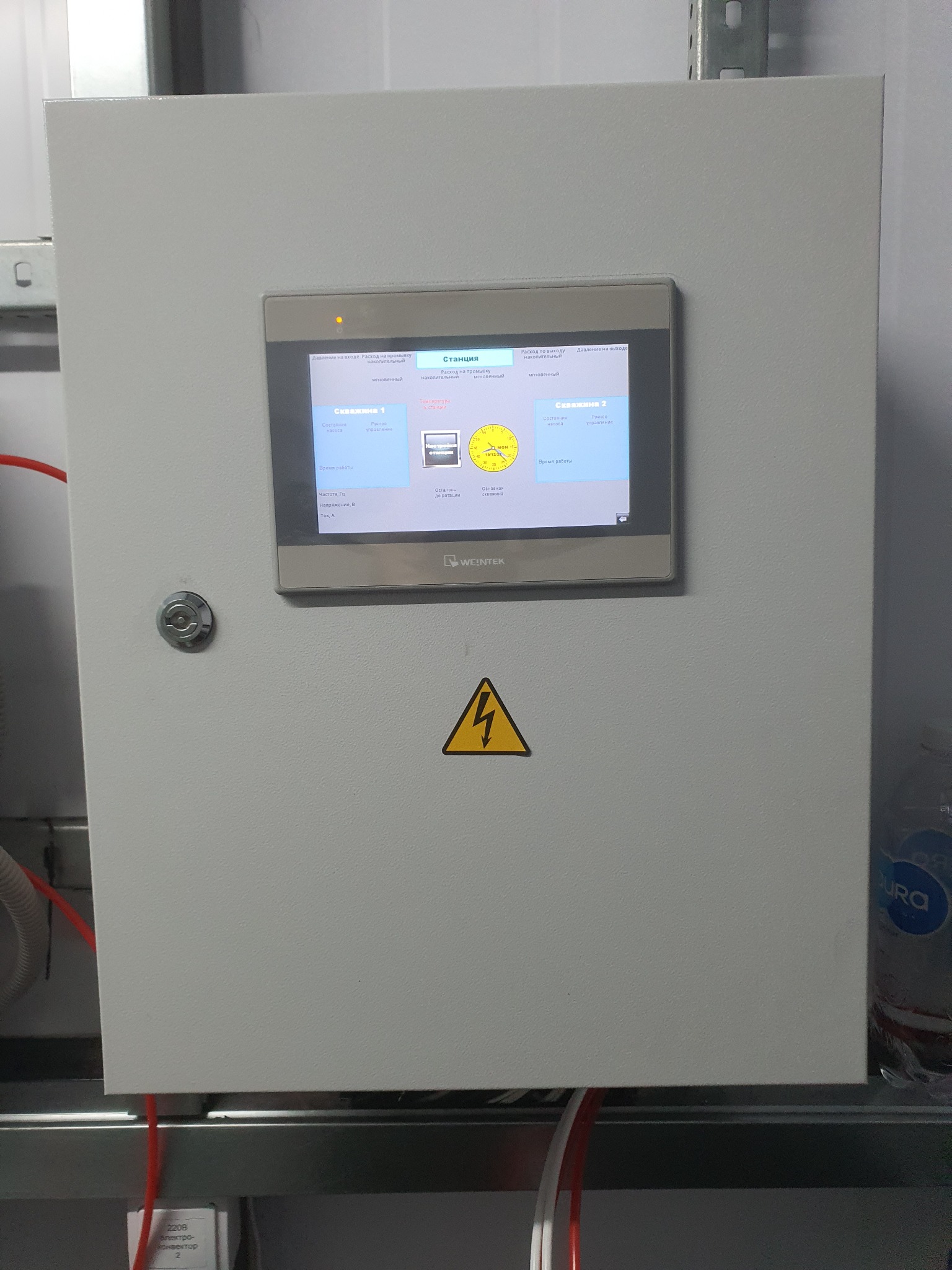 Szara szafa sterownicza z panelem dotykowym Weintek wyświetlającym schemat stacji z dwoma studniami, żółty znak ostrzegawczy z symbolem błyskawicy
