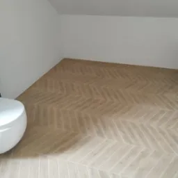 Ułożona jasna podłoga z płytek imitujących drewno w jodełkę w łazience na poddaszu, widoczny fragment białej toalety.