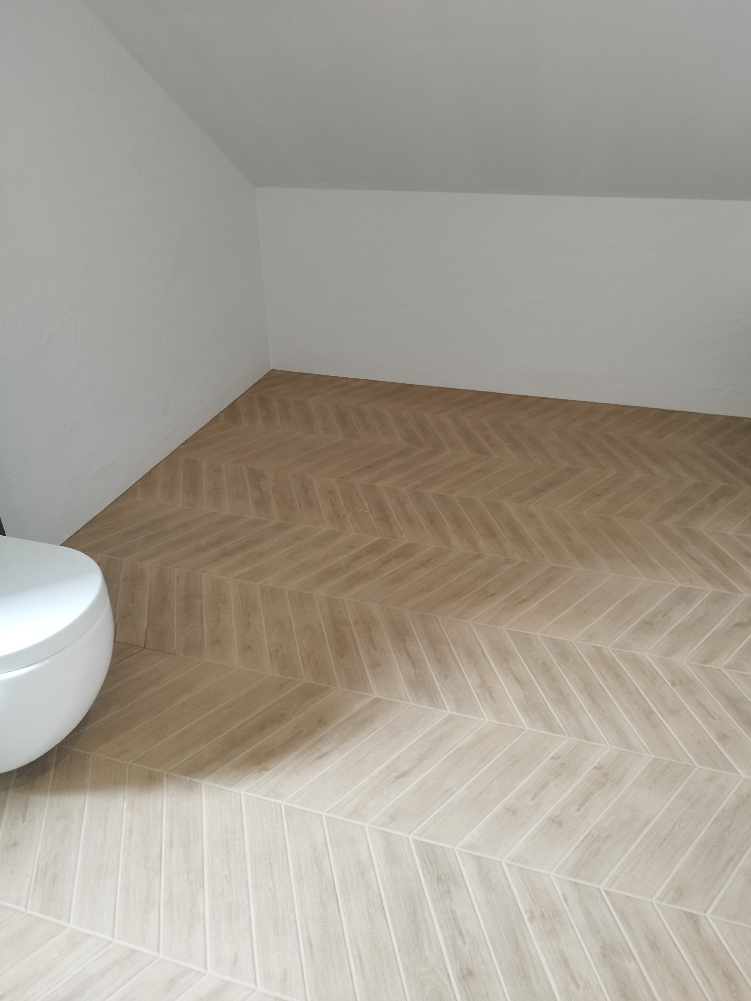 Ułożona jasna podłoga z płytek imitujących drewno w jodełkę w łazience na poddaszu, widoczny fragment białej toalety.