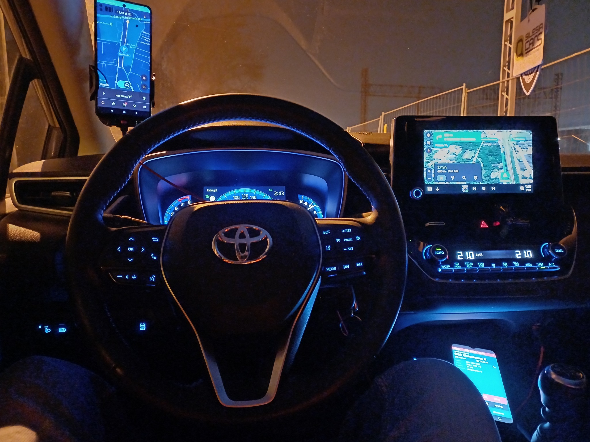 Wnętrze samochodu Toyota z włączonymi nawigacjami na dwóch ekranach: smartfonie na uchwycie i wbudowanym ekranie dotykowym, widoczne logo Silesia Cars na zewnątrz.