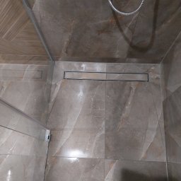 Czesław Boratyn Loft&Design usługi remontowe - Wykończona kabina prysznicowa z odpływem liniowym i marmurkowymi płytkami w odcieniach beżu i brązu, widok z góry, fragment buta w dolnej części kadru.