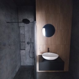 Czesław Boratyn Loft&Design usługi remontowe - Nowoczesna łazienka w odcieniach szarości z drewnianym akcentem: kabina prysznicowa typu walk-in, umywalka nablatowa i okrągłe lustro na tle pionowych paneli.