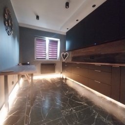 Czesław Boratyn Loft&Design usługi remontowe - Nowoczesna kuchnia z czarnymi szafkami, marmurową podłogą i podświetleniem LED. Na ścianie zegar i dekoracja w kształcie serca. Okno z żaluzjami.