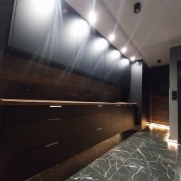 Czesław Boratyn Loft&Design usługi remontowe - Nowoczesna, ciemna kuchnia z marmurową podłogą i oświetleniem punktowym. Długi blat z drewnianym akcentem i minimalistyczne szafki tworzą elegancką przestrzeń.