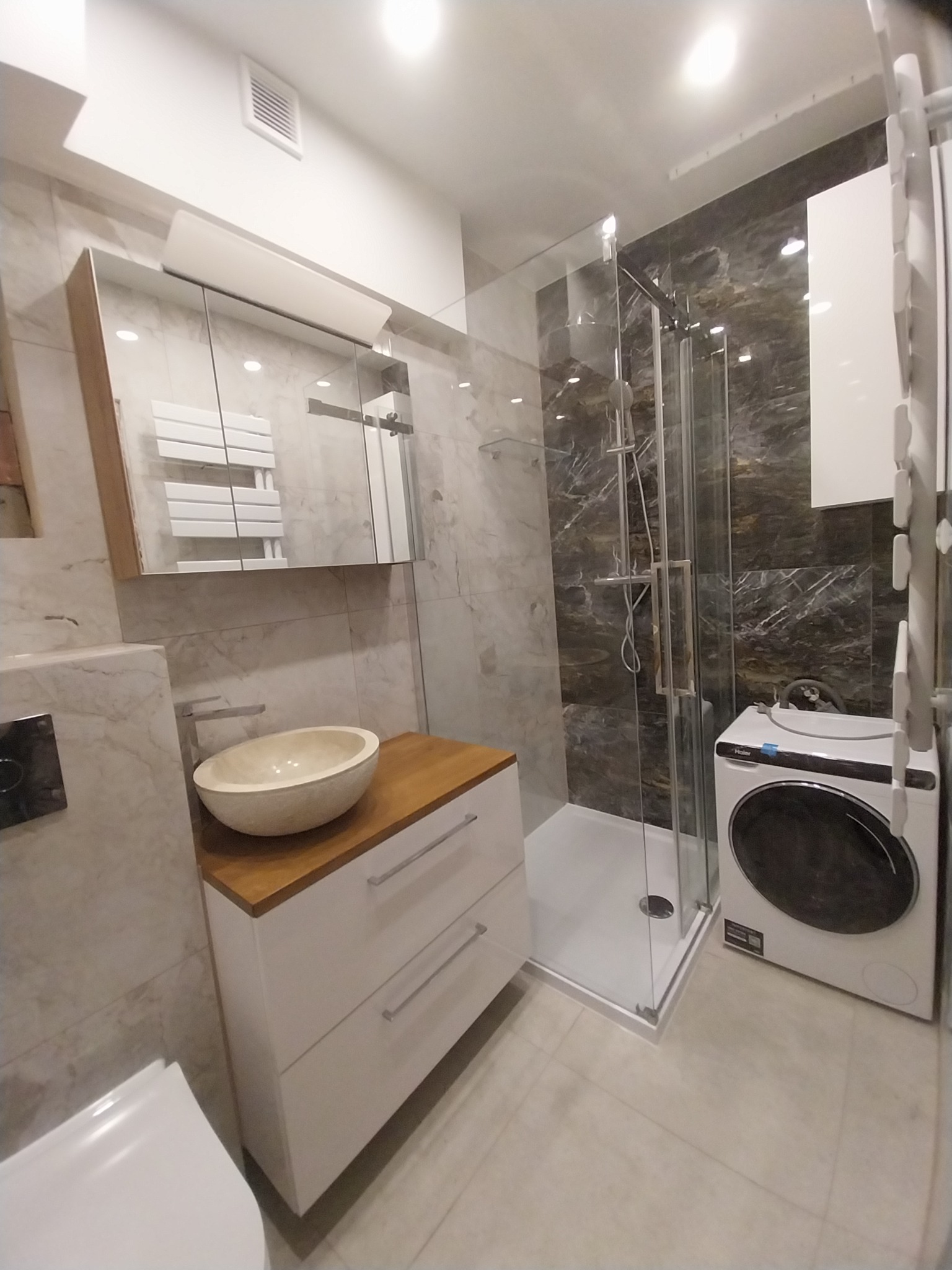 Nowoczesna łazienka z kabiną prysznicową typu walk-in, umywalką nablatową w kształcie misy, szafką z dwiema szufladami, pralką i grzejnikiem drabinkowym w szafce z lustrem.