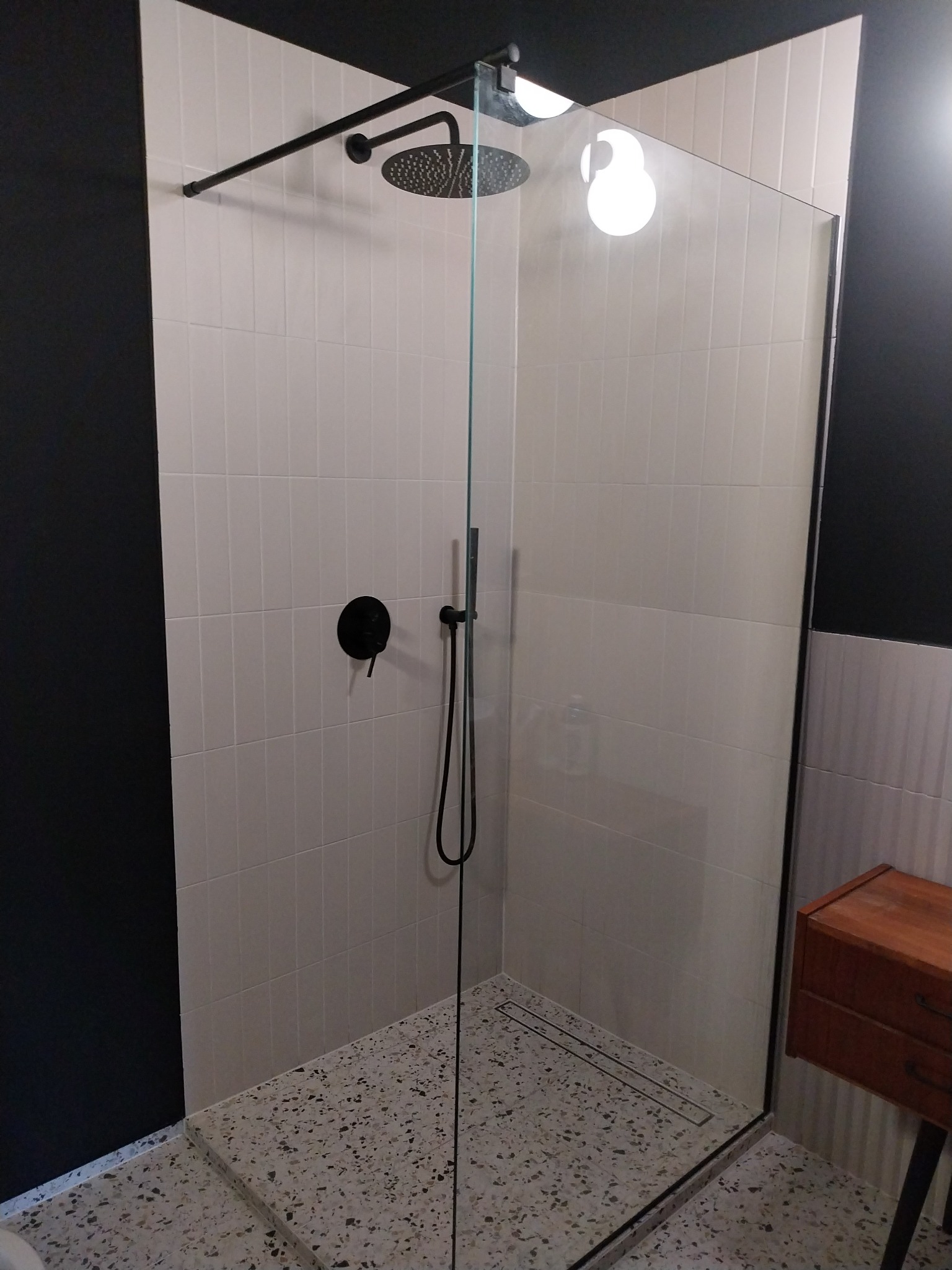 Kabina prysznicowa walk-in z czarną armaturą, białe płytki ułożone pionowo, podłoga lastryko, czarna ściana w tle, widoczna część drewnianej szafki.